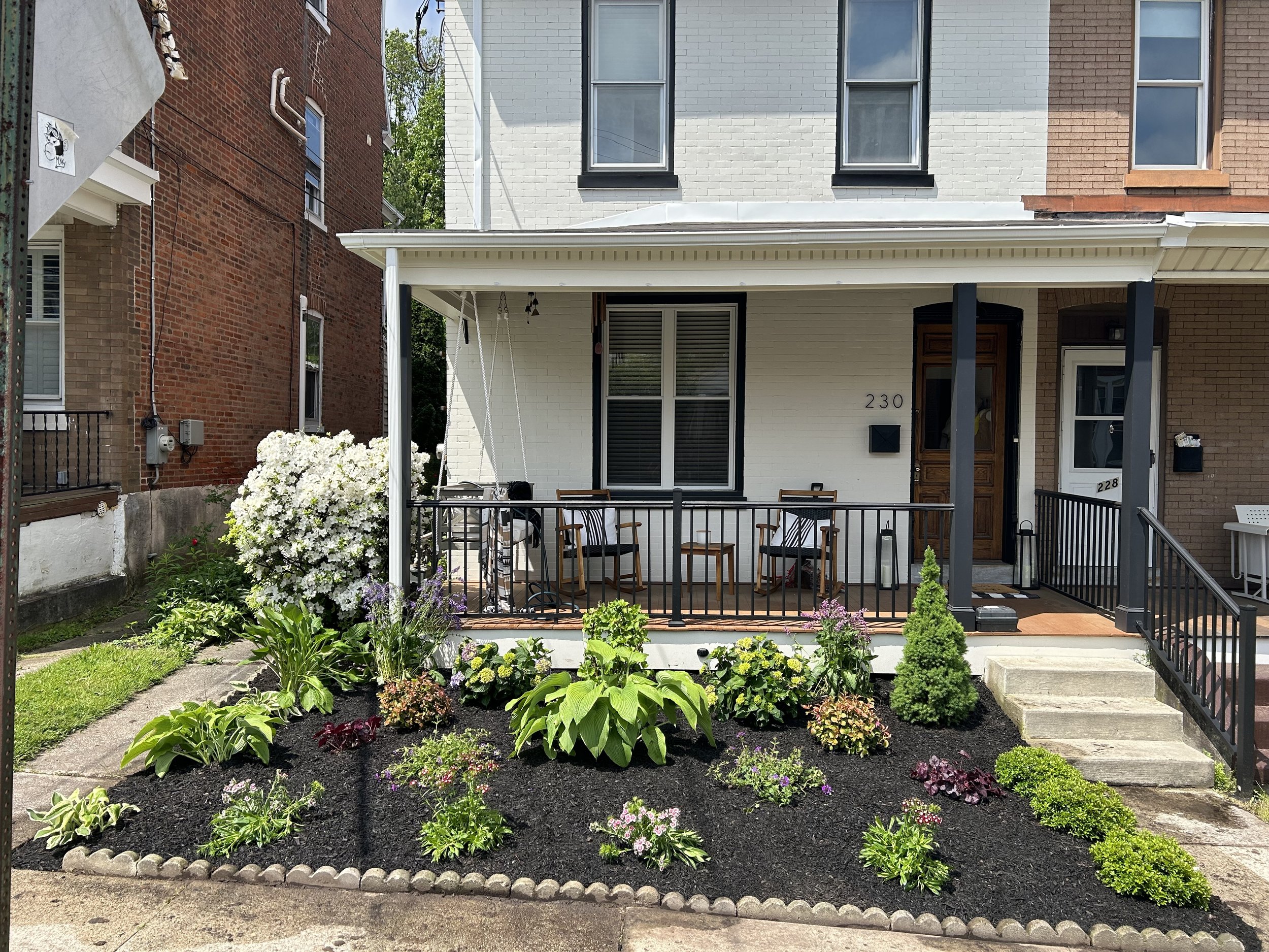 A white brick house with a small porch and landscaped front yard. The porch has a black railing, two wooden chairs, and a white door with the number 230. The front yard features various green plants and shrubs with mulch, bordered by a concrete sidew