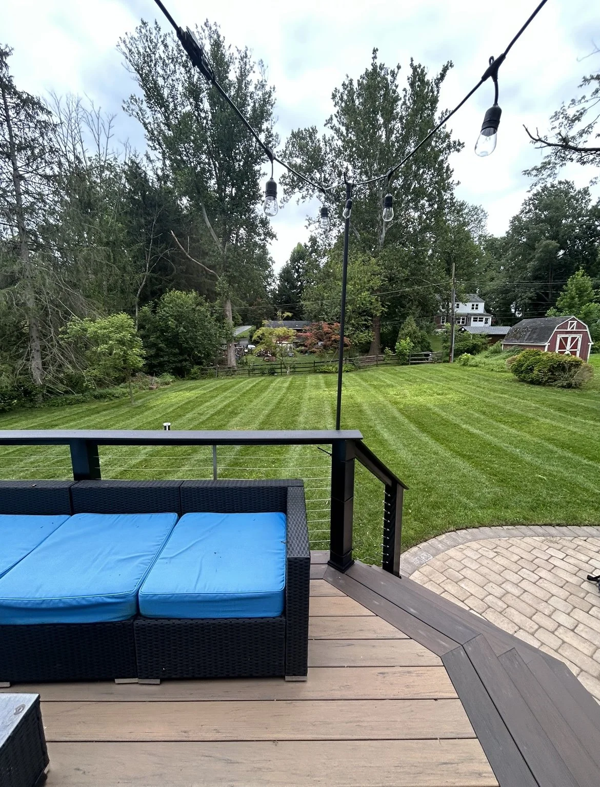 Backyard view with wooden deck, blue cushioned patio furniture, hanging string lights, neatly mowed lawn, and surrounding trees.