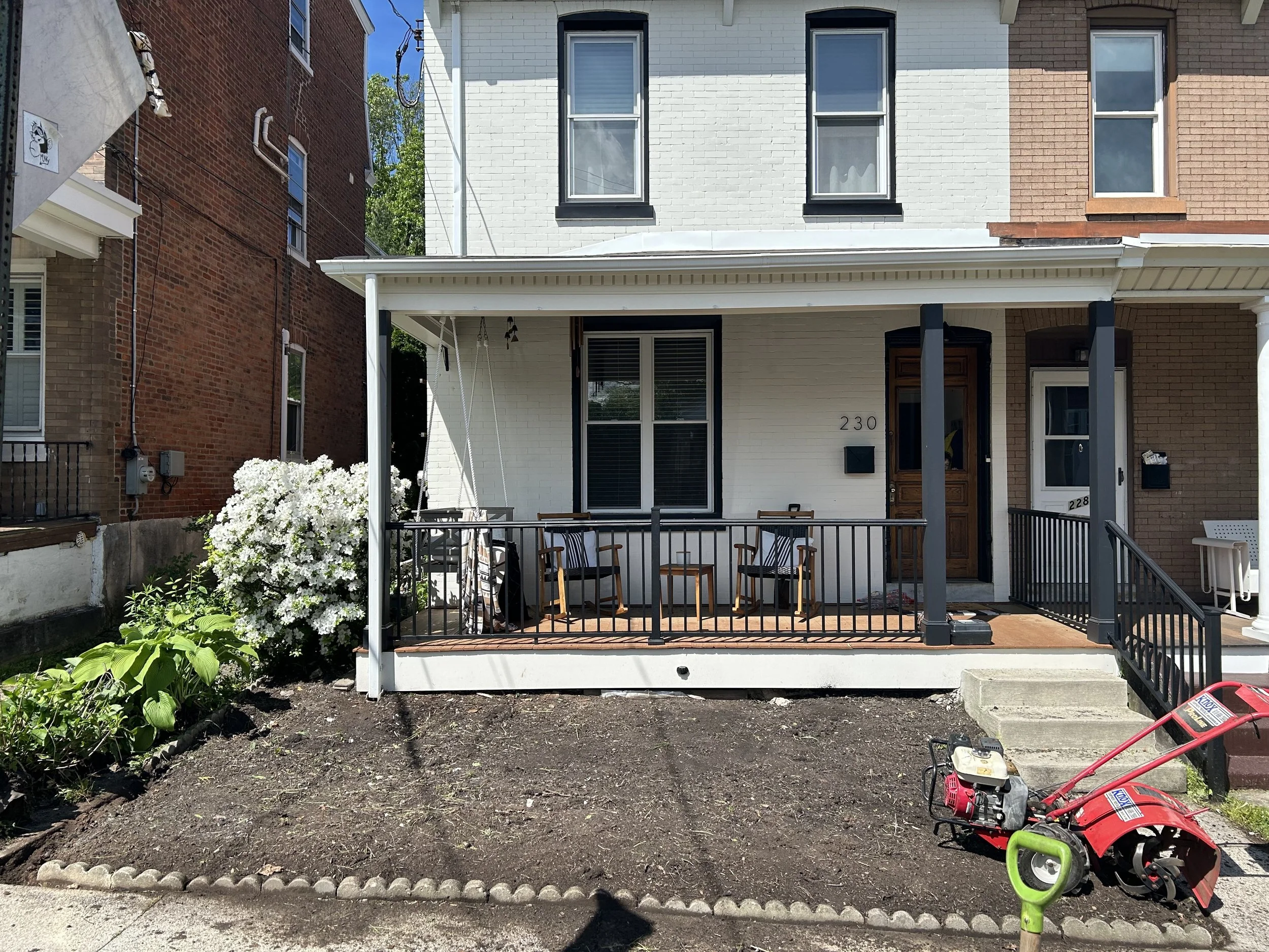 Front view of a townhouse with a small porch, chairs, railing, and a garden area being prepared for planting. A red tiller and a green shovel are in the dirt area. There's a white brick wall, black window frames, and a wooden door. Adjacent houses an