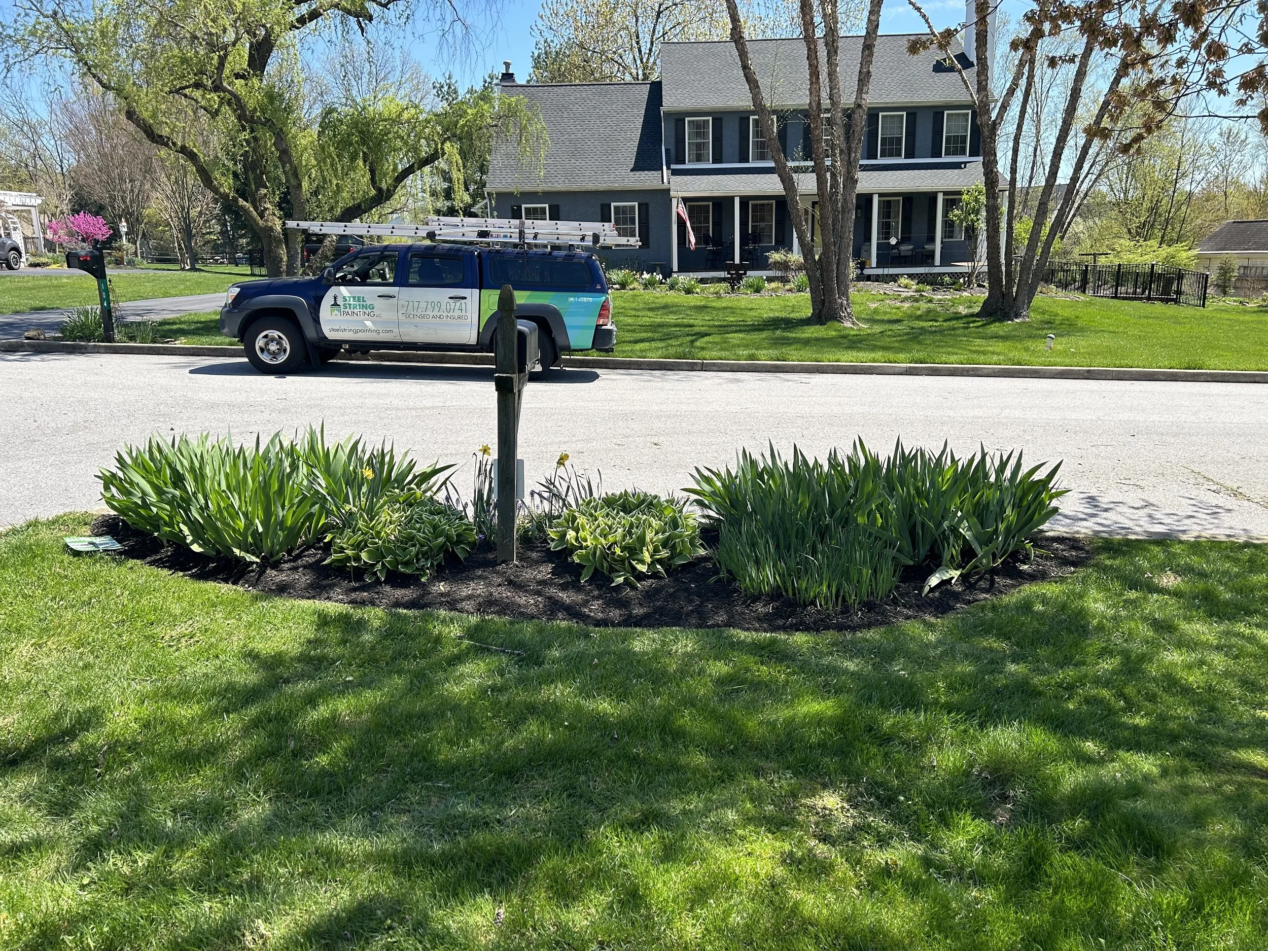A suburban street with a landscaped lawn, featuring green plants and shrubs surrounding a wooden post. A blue pickup truck with a ladder on top is parked on the road in front of a two-story house with a porch and American flag. Trees and a neatly tri