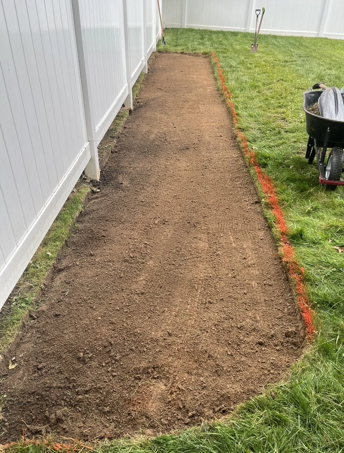 Dug-out pathway with soil bordered by grass and white fence, garden tools, and wheelbarrow.