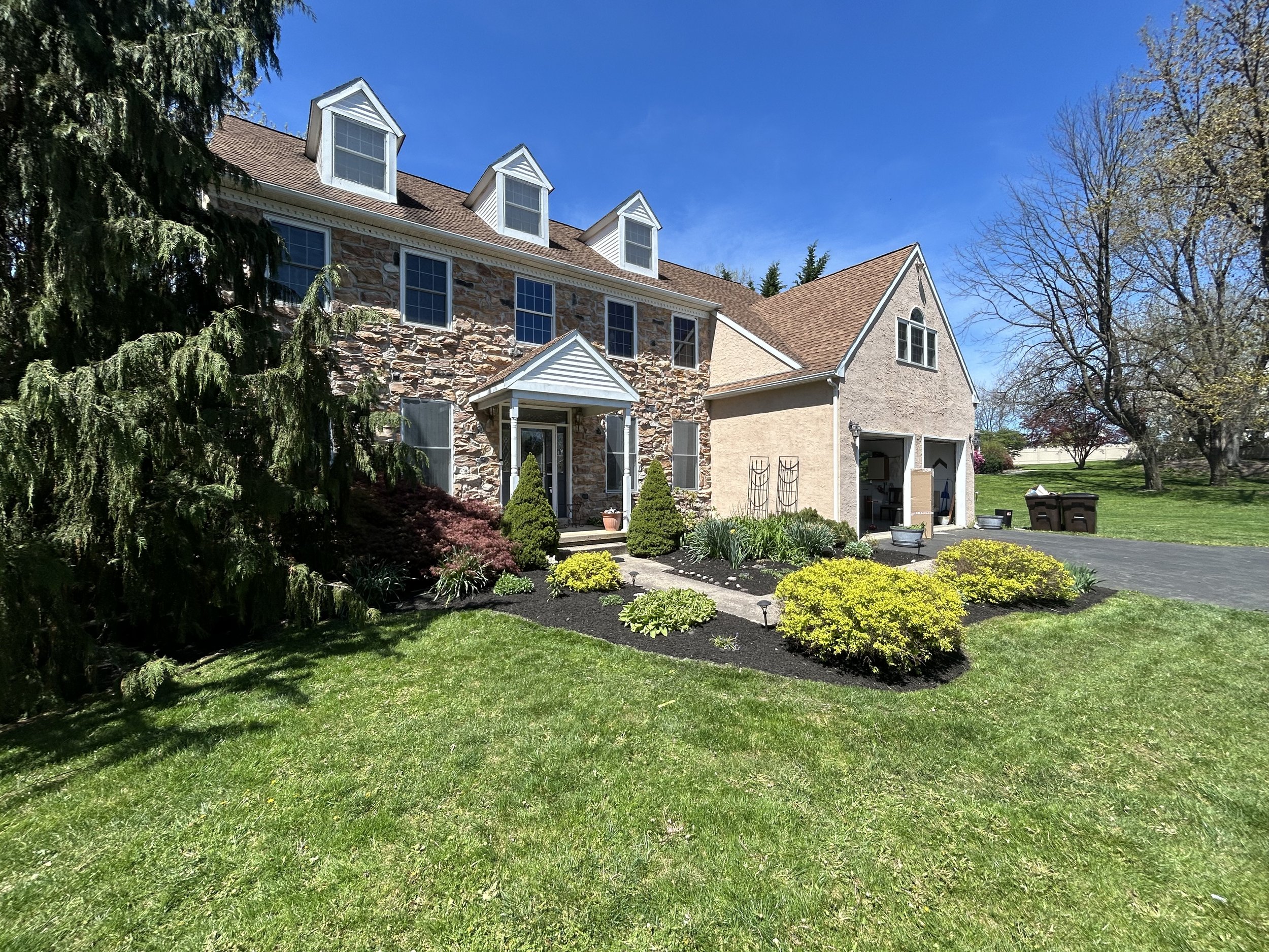 Stone house with dormer windows, manicured lawn, and driveway on a sunny day.