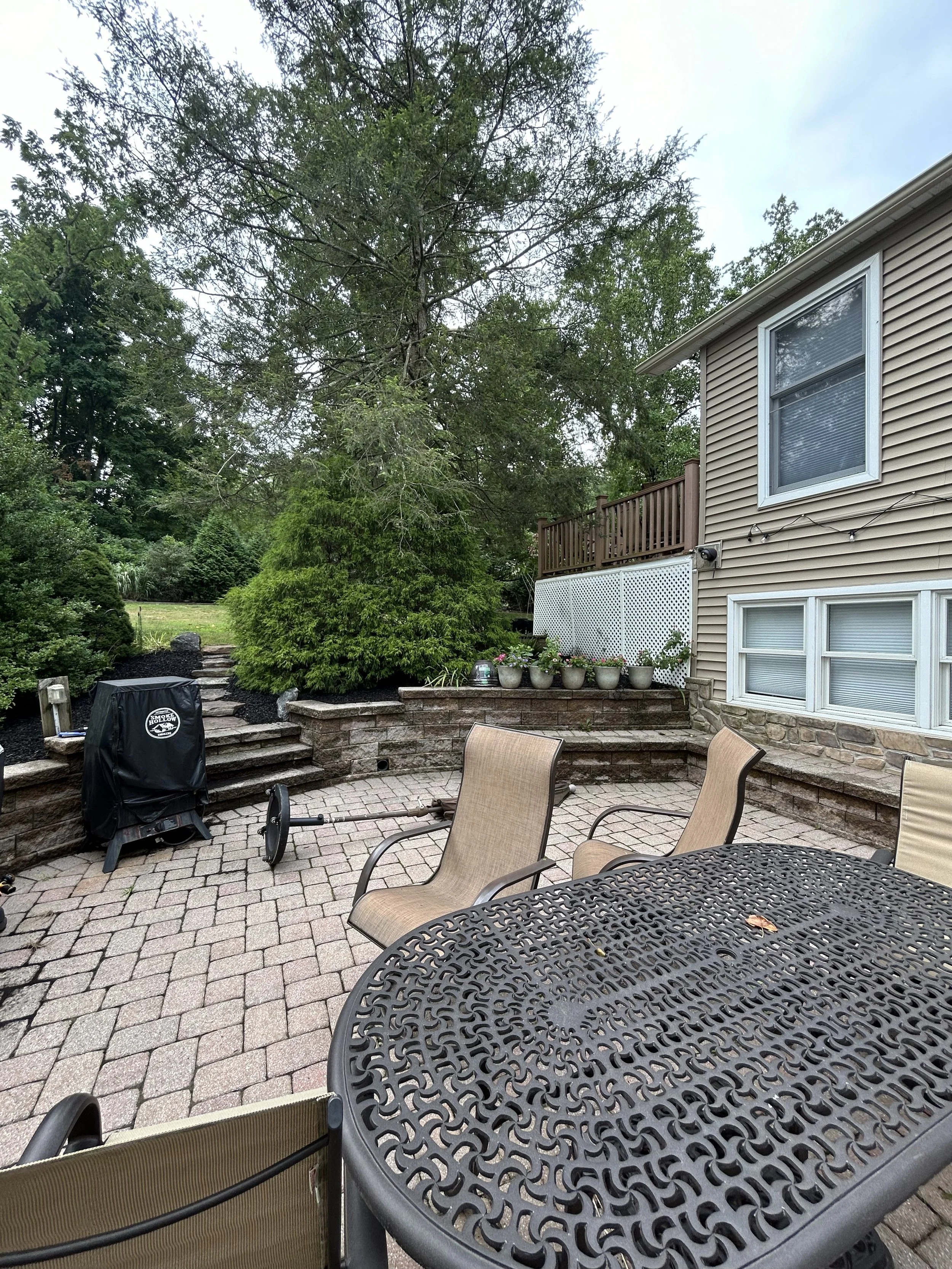 Patio with metal table, chairs, grill covered with black cover, stone steps, potted plants, large trees, and tan house with windows.