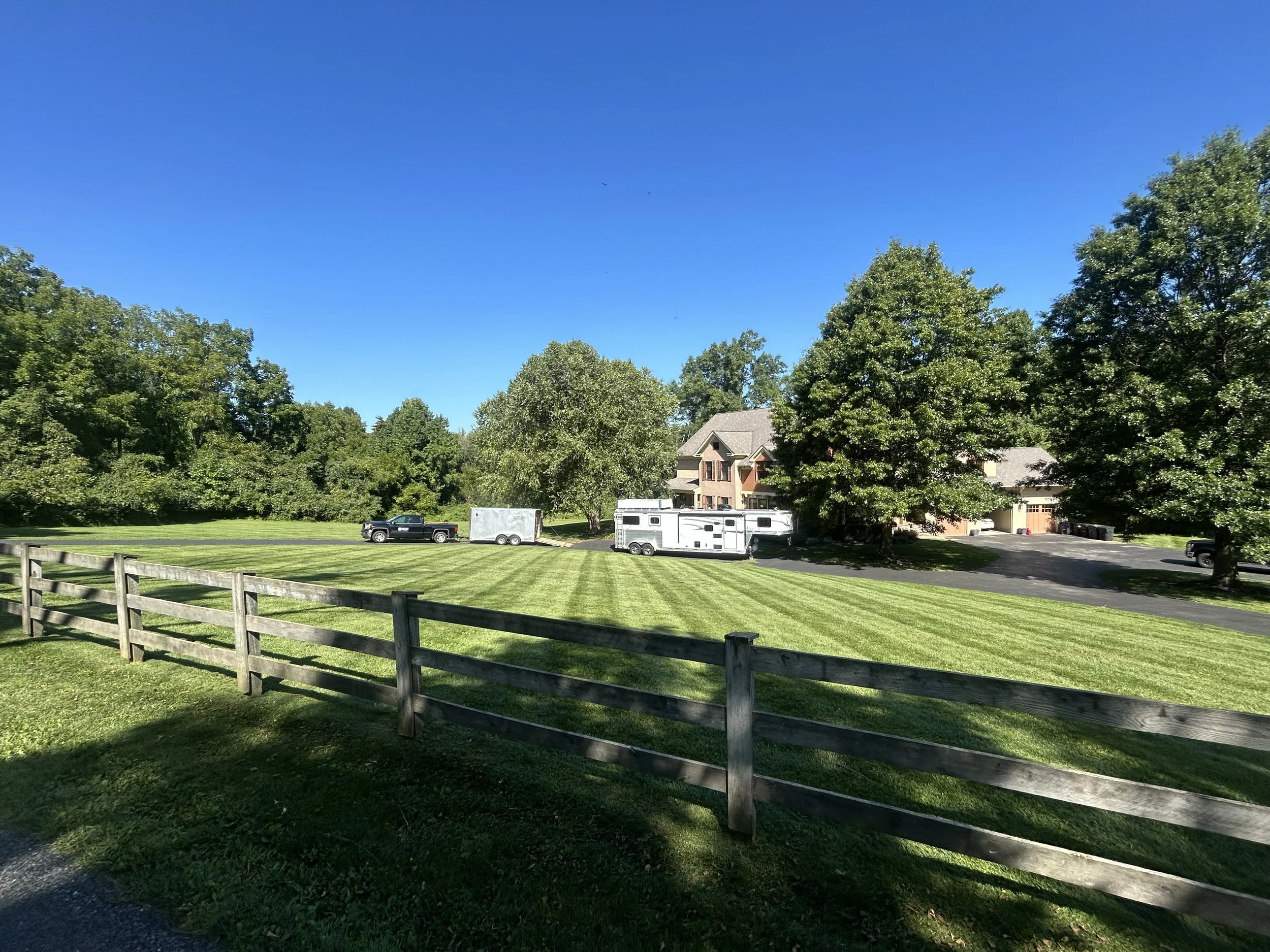 Rural property with a large house, grassy yard, and parked vehicles, including a truck with a trailer, set against a backdrop of trees.