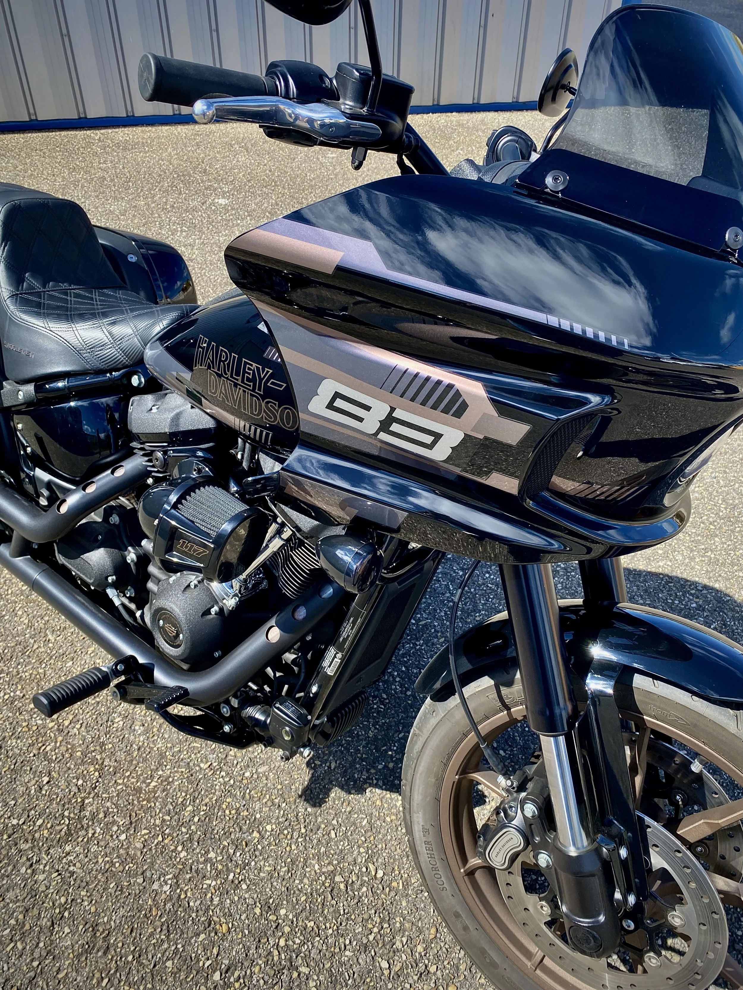 Une moto Harley-Davidson noire avec des détails gris et bronzes, vue en plein jour, avec un pare-brise et des pneus de grande taille, stationnée sur une surface asphaltée.