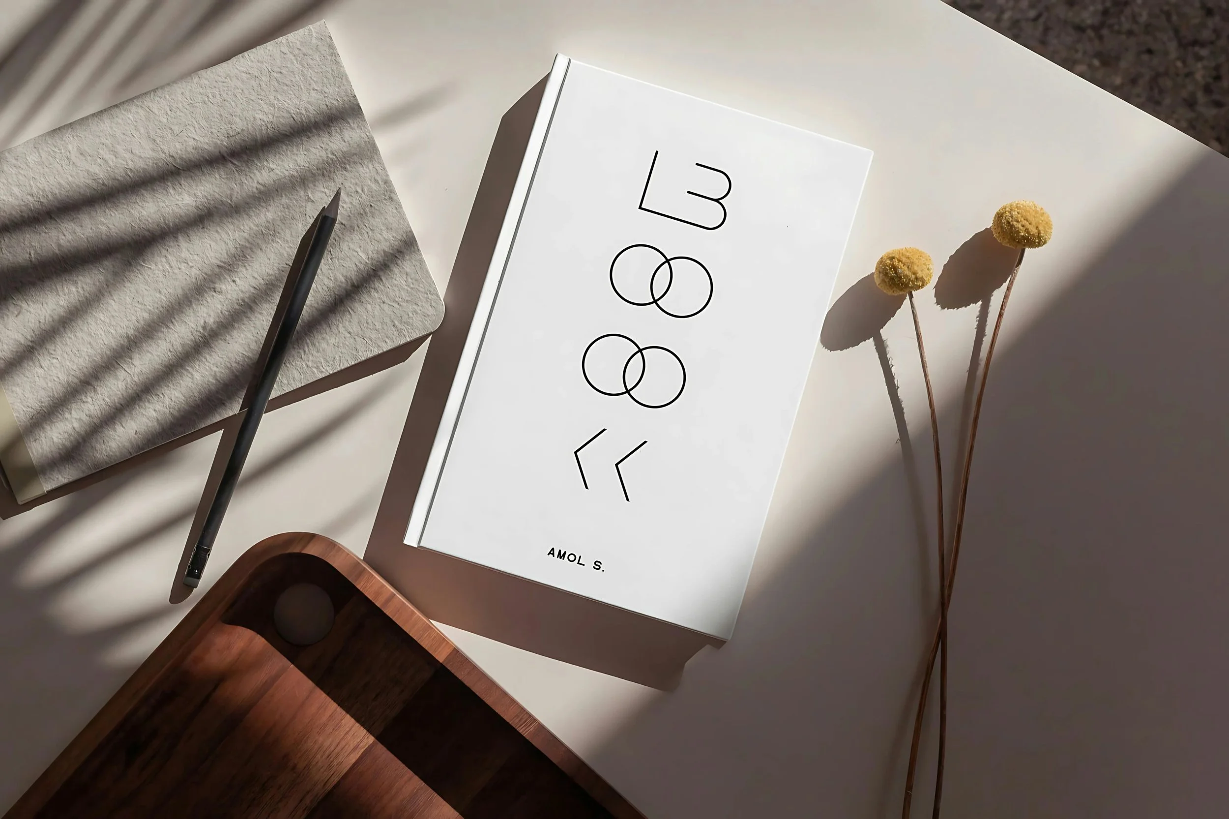 Minimalist white book cover with modern black typography reading "BOOK," displayed on a neutral desk with a pencil, notebook, and dried yellow Billy Button flowers.
