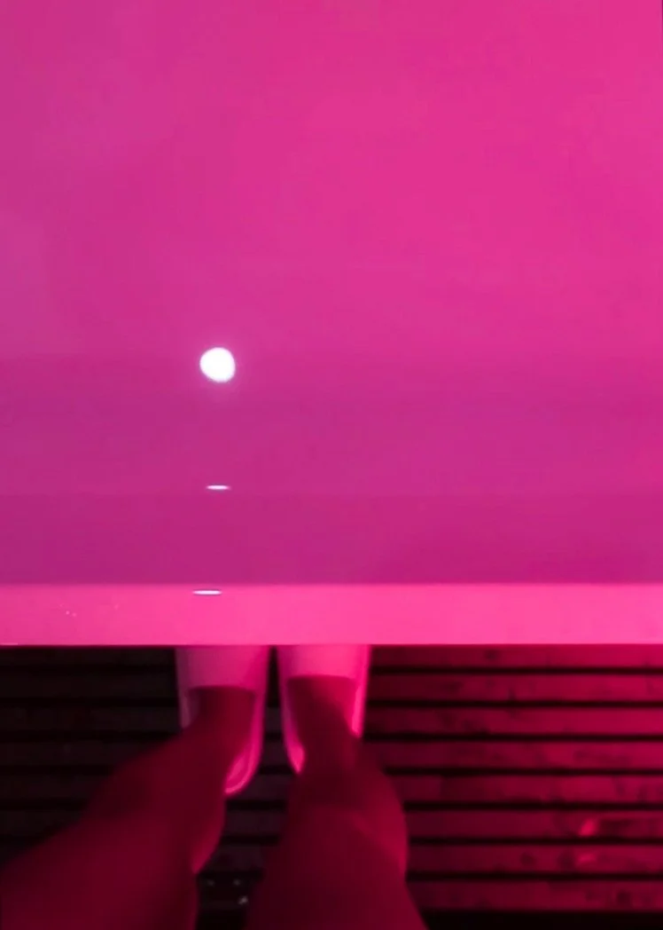 Person standing barefoot on a wooden deck, looking down at a pink-lit platform with a bright white light and reflections on the surface.