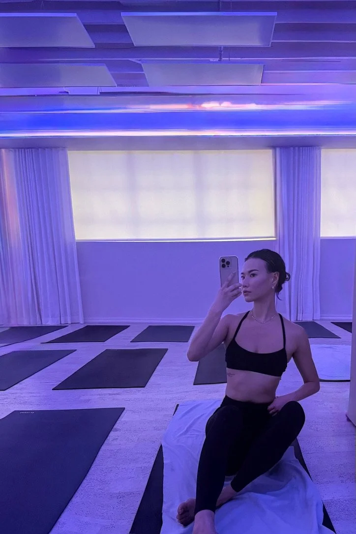 A woman taking a selfie in a yoga studio with purple lighting, sitting on a yoga mat, surrounded by several other yoga mats in the room.