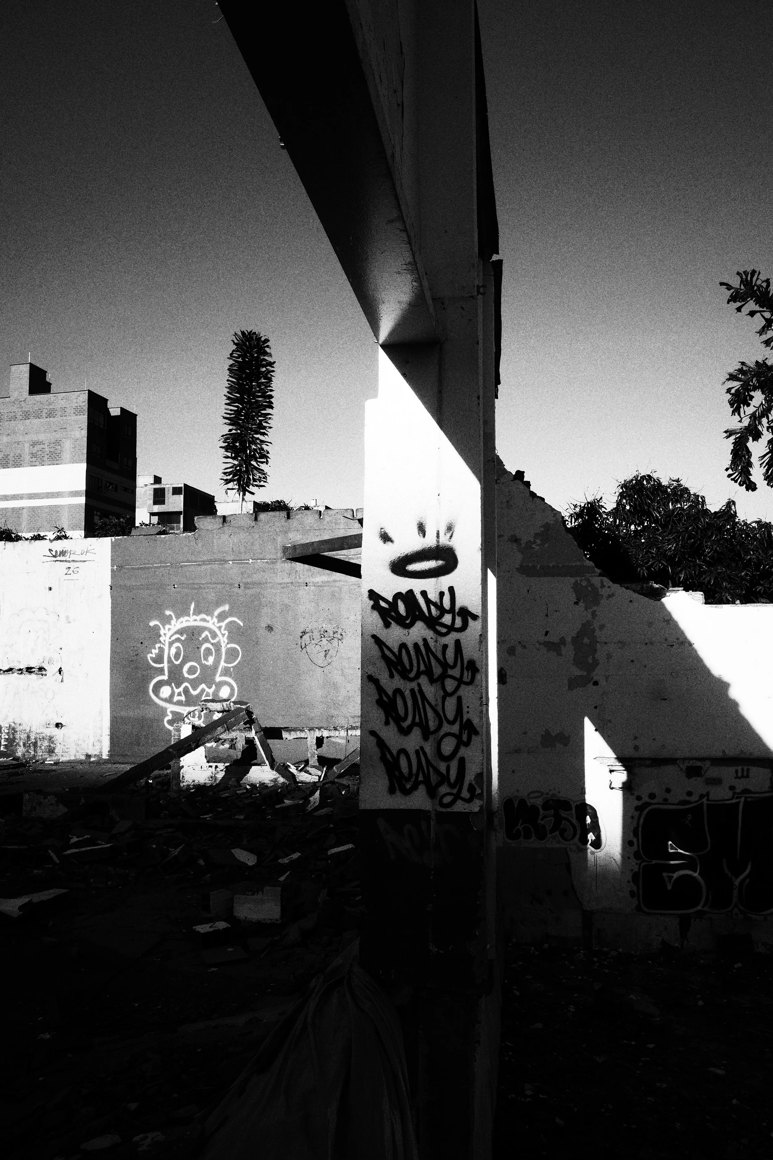 Black and white photo of a ruined building interior in Medellín, where a single concrete beam casts deep shadows across graffiti-covered walls and scattered debris.