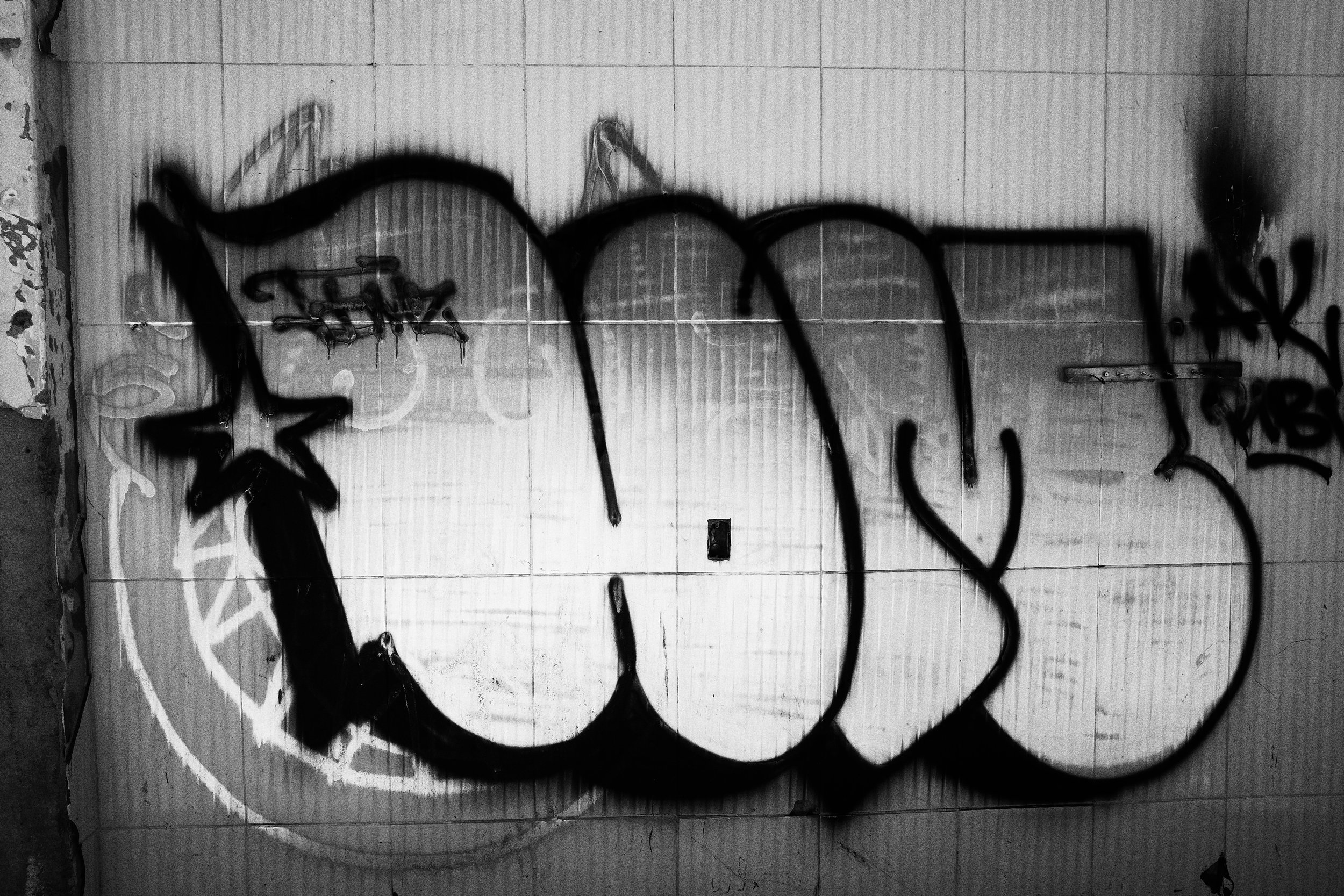 Black and white photo of oversized graffiti painted on a tiled interior wall of a demolished building in Medellín, the soft spray paint edges contrasting with the rigid grid of the tiles.