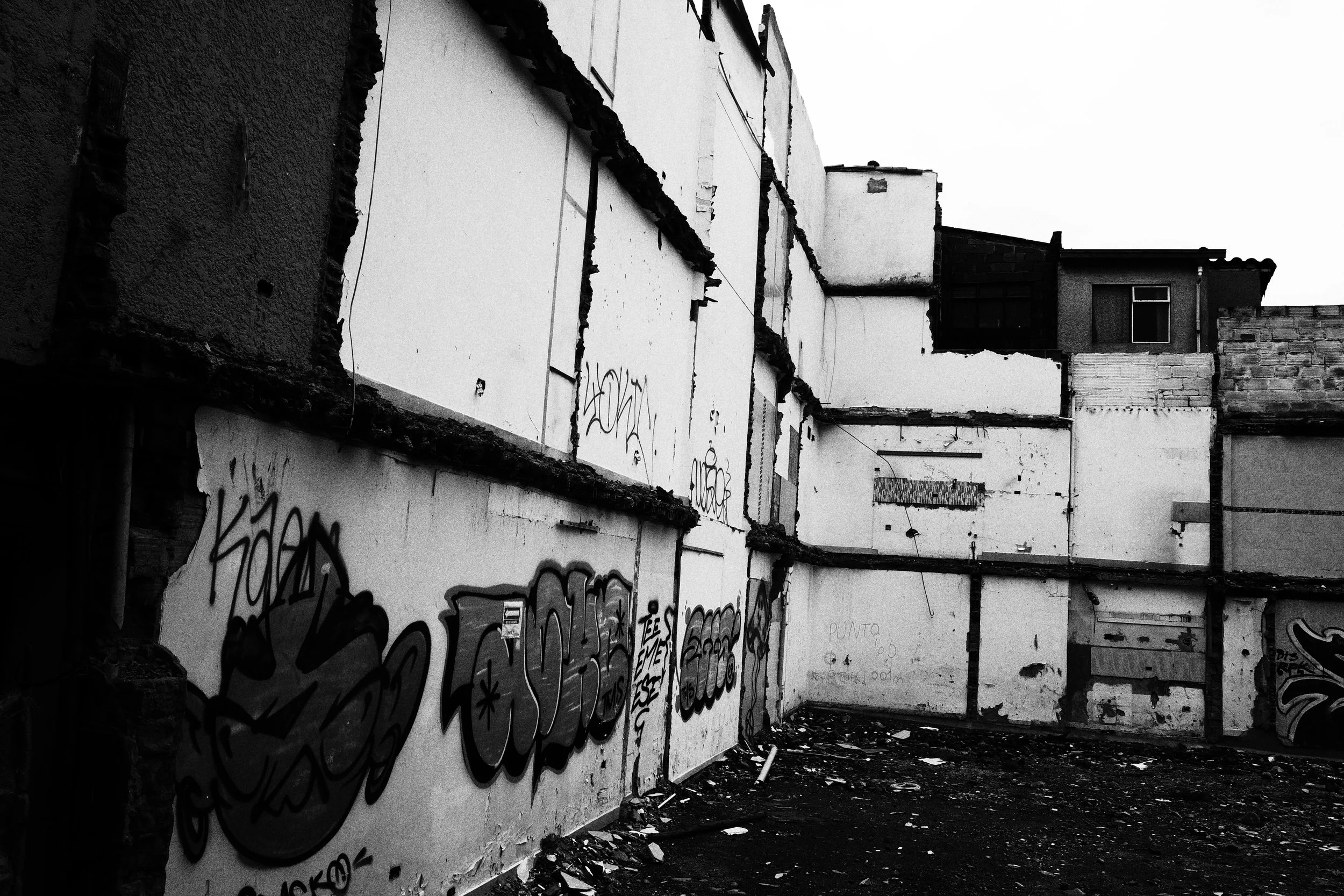 Black and white photo of the exposed interior walls of demolished homes in Medellín, covered in graffiti and opening onto an empty lot scattered with debris.
