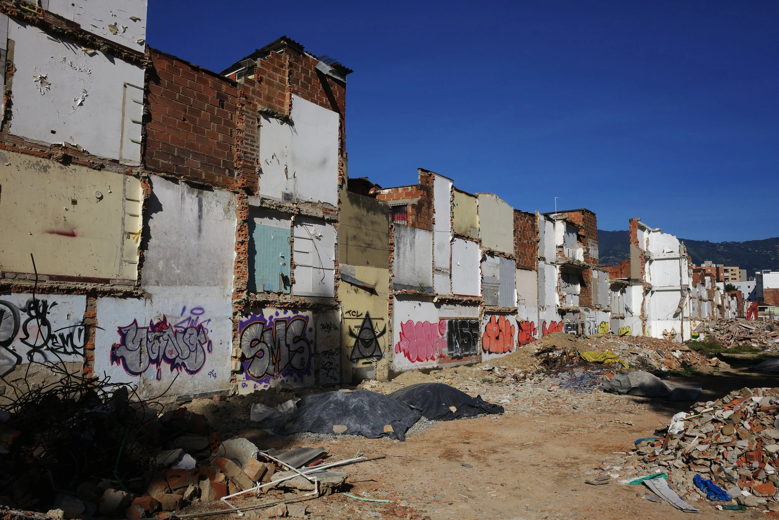 Color photo of a long row of demolished homes in Medellín, with exposed brick walls, colorful graffiti, and piles of rubble stretching toward the mountains in the distance.