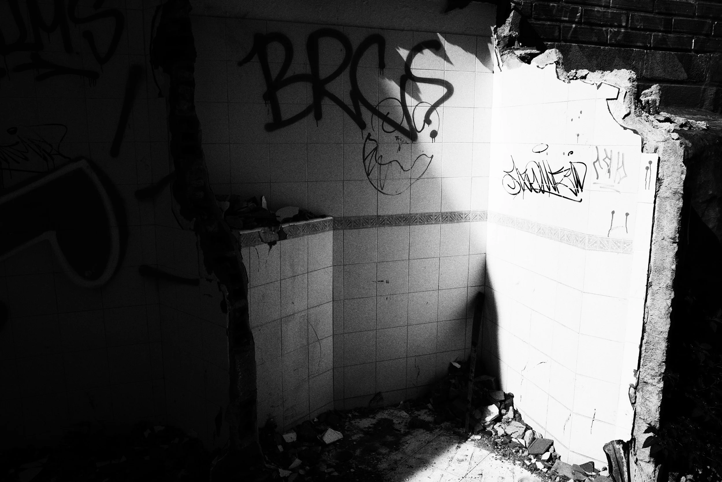 Black and white photo of a partially destroyed tiled room in Medellín, where harsh sunlight cuts across graffiti-covered walls and broken masonry on the floor.