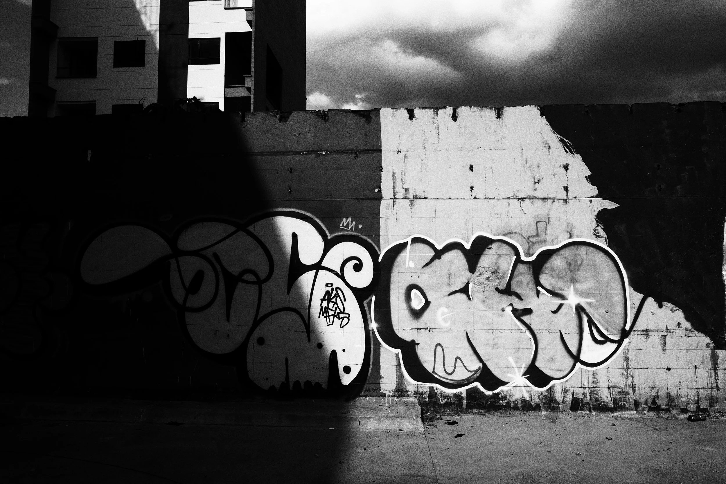 Black and white photo of large graffiti tags painted across a partially demolished wall in Medellín, divided sharply by shadow beneath dark storm clouds and apartment buildings.