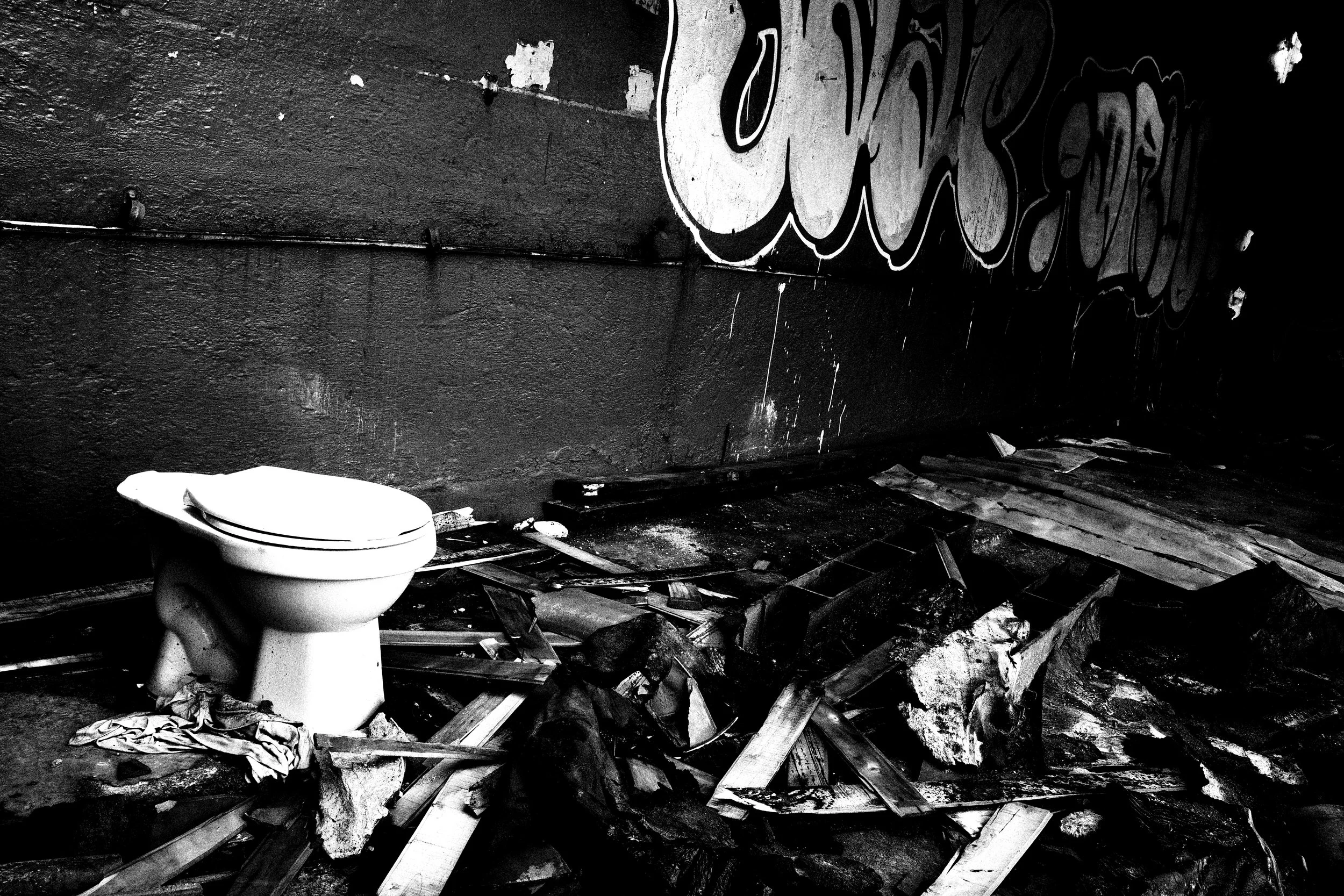 Black and white photo of a white toilet standing alone among broken wood, rubble, and graffiti inside a dark abandoned structure in Medellín.