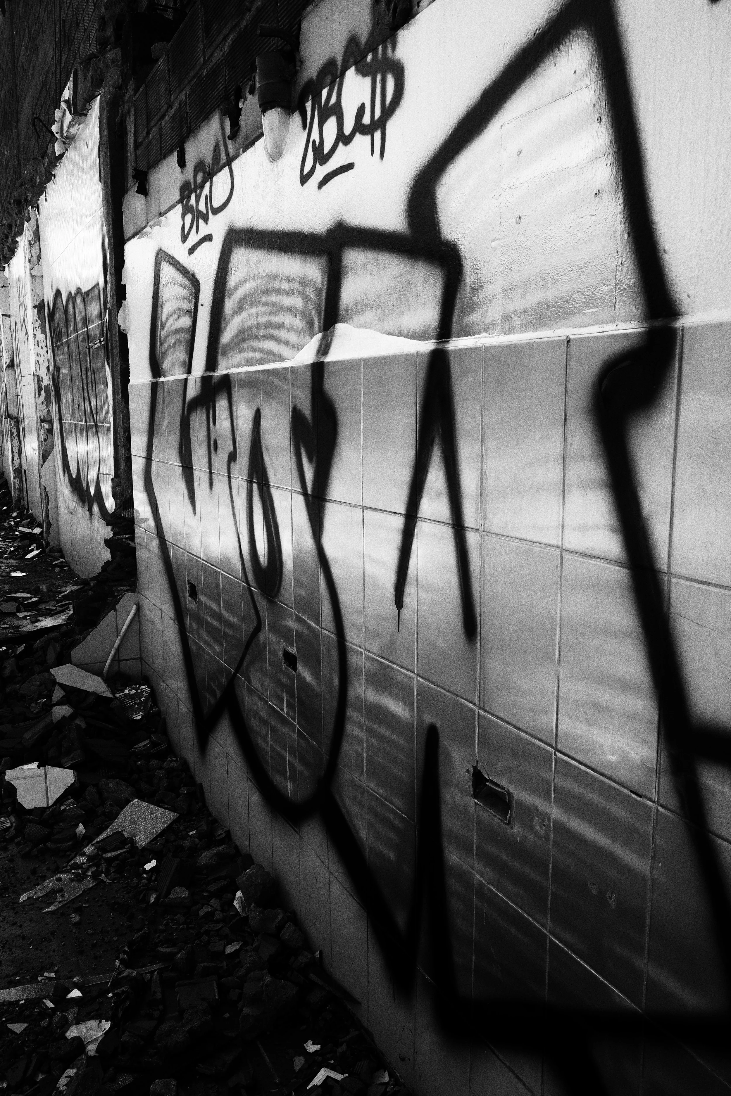 Black and white photo taken along a narrow corridor inside a demolished building in Medellín, with large graffiti tags stretching across tiled walls above piles of broken rubble.
