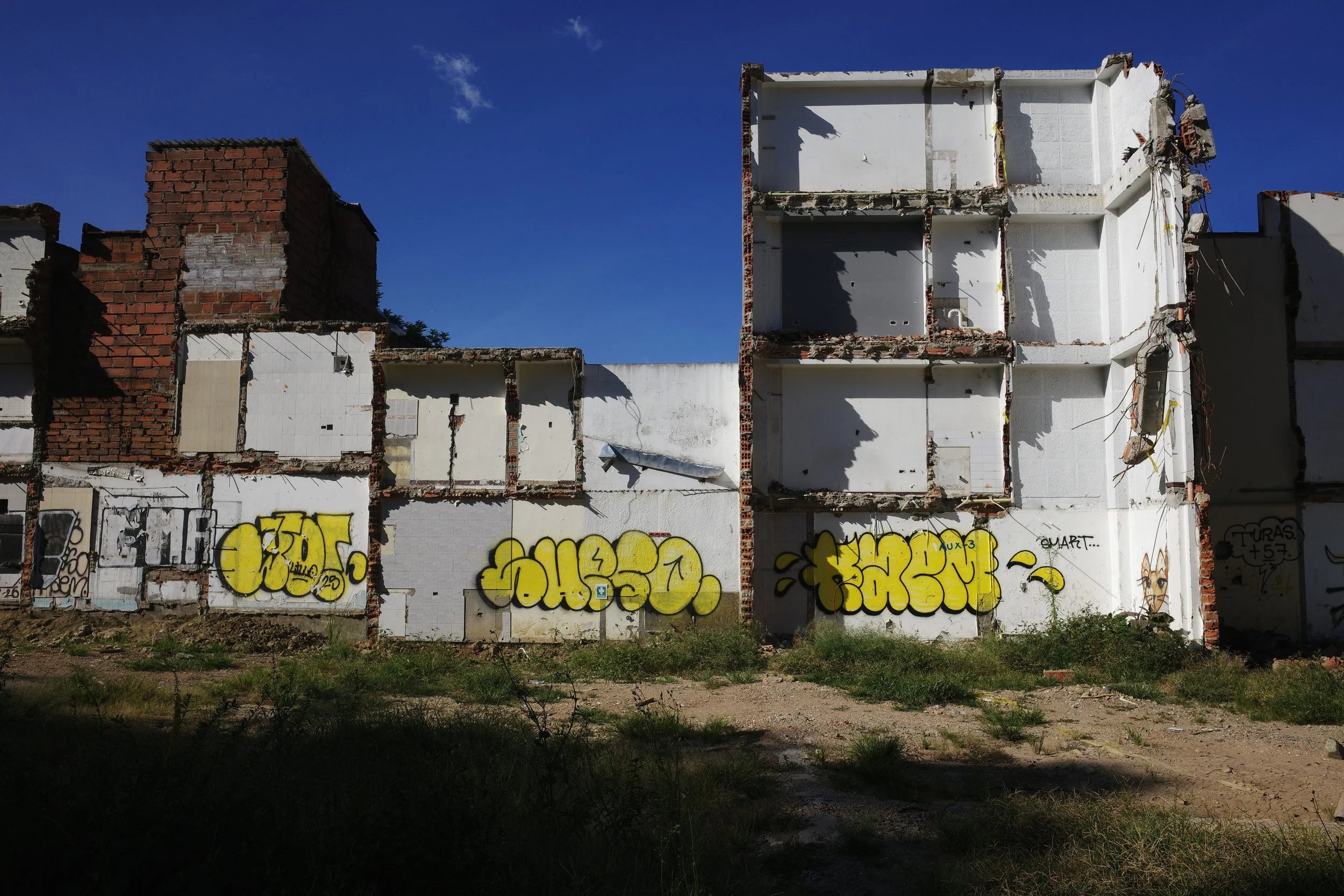 Color photo of partially demolished apartment buildings in Medellín, their exposed white interiors and yellow graffiti standing beneath a deep blue sky.