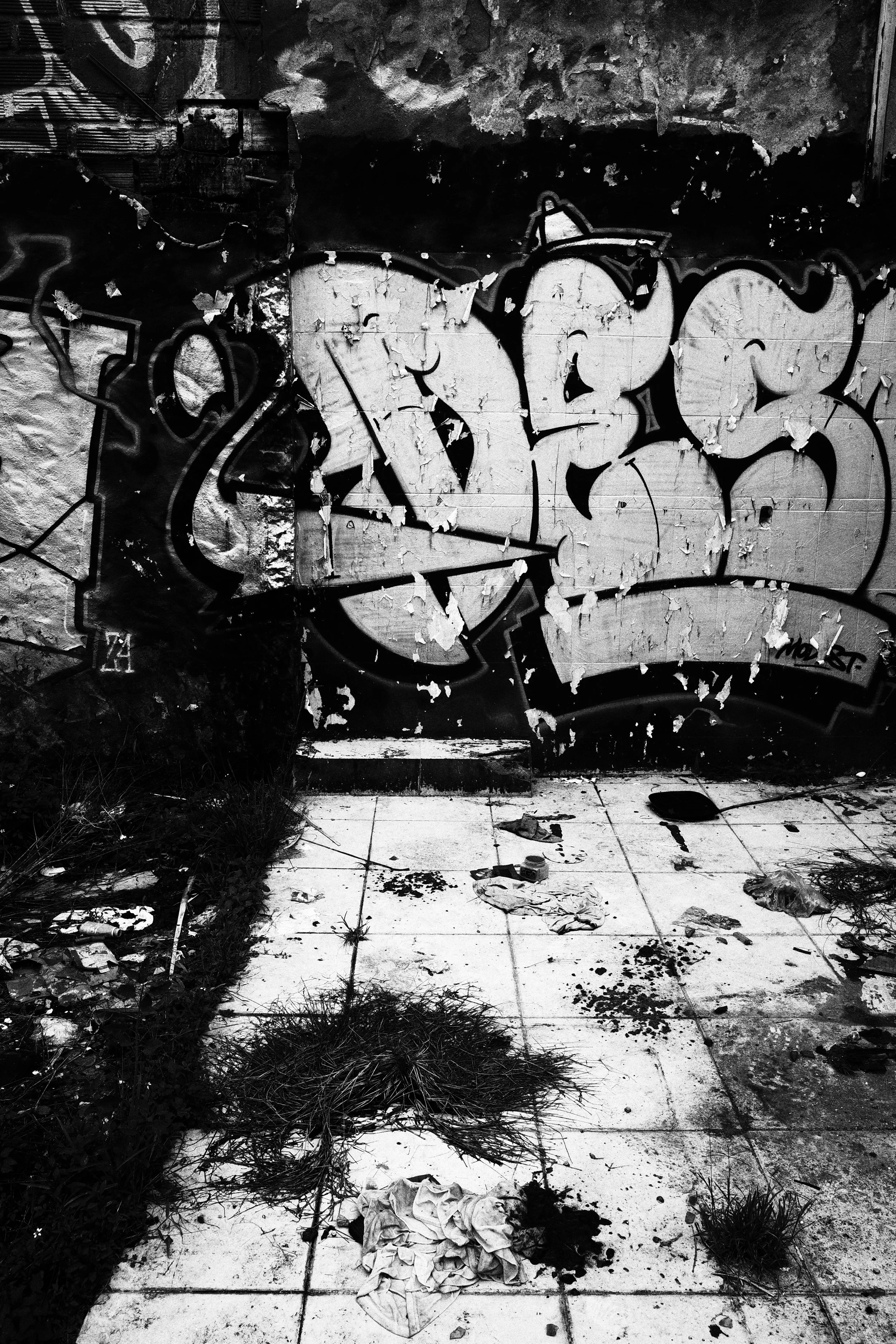 Black and white photo of peeling graffiti covering the tiled wall of an abandoned room in Medellín, with weeds and scraps of cloth scattered across the dirty floor.
