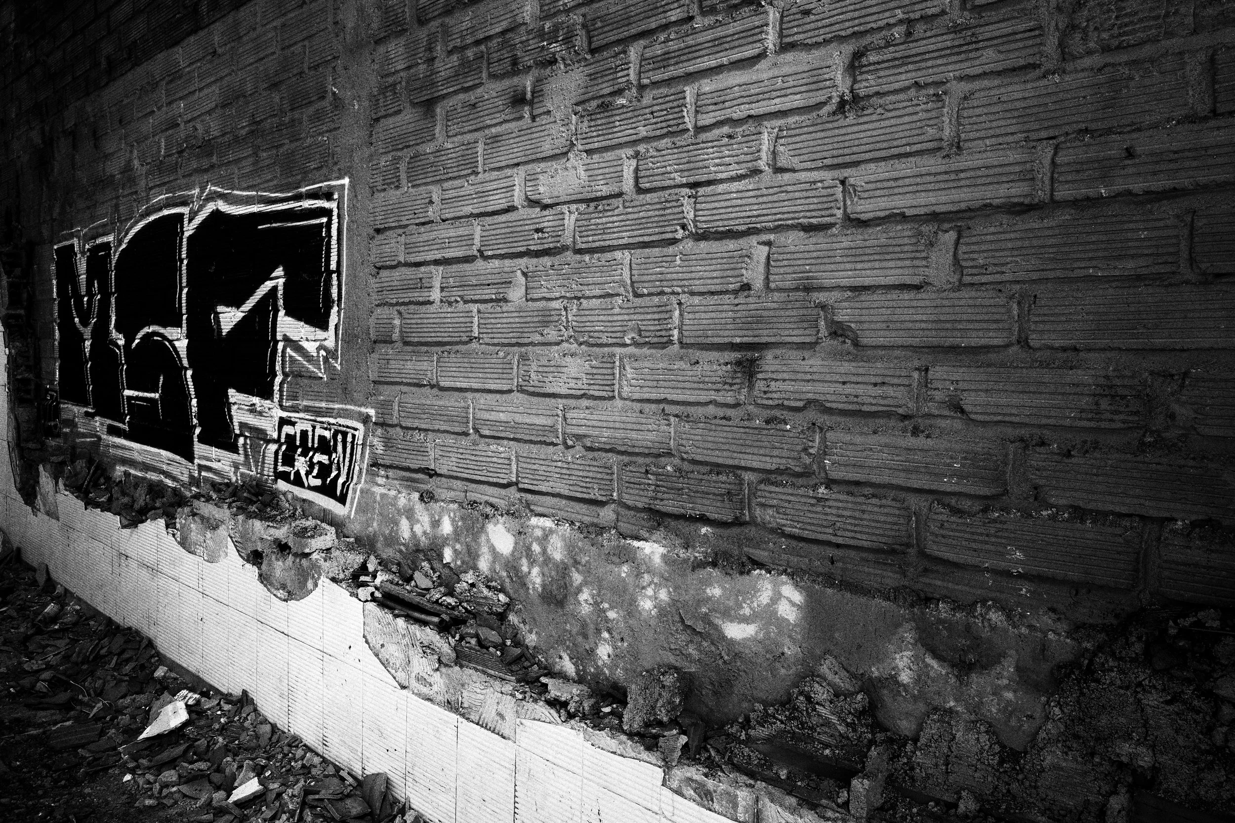 Black and white photo of graffiti sprayed across a damaged brick wall inside a partially demolished building in Medellín, with broken tiles and rubble scattered along the floor.