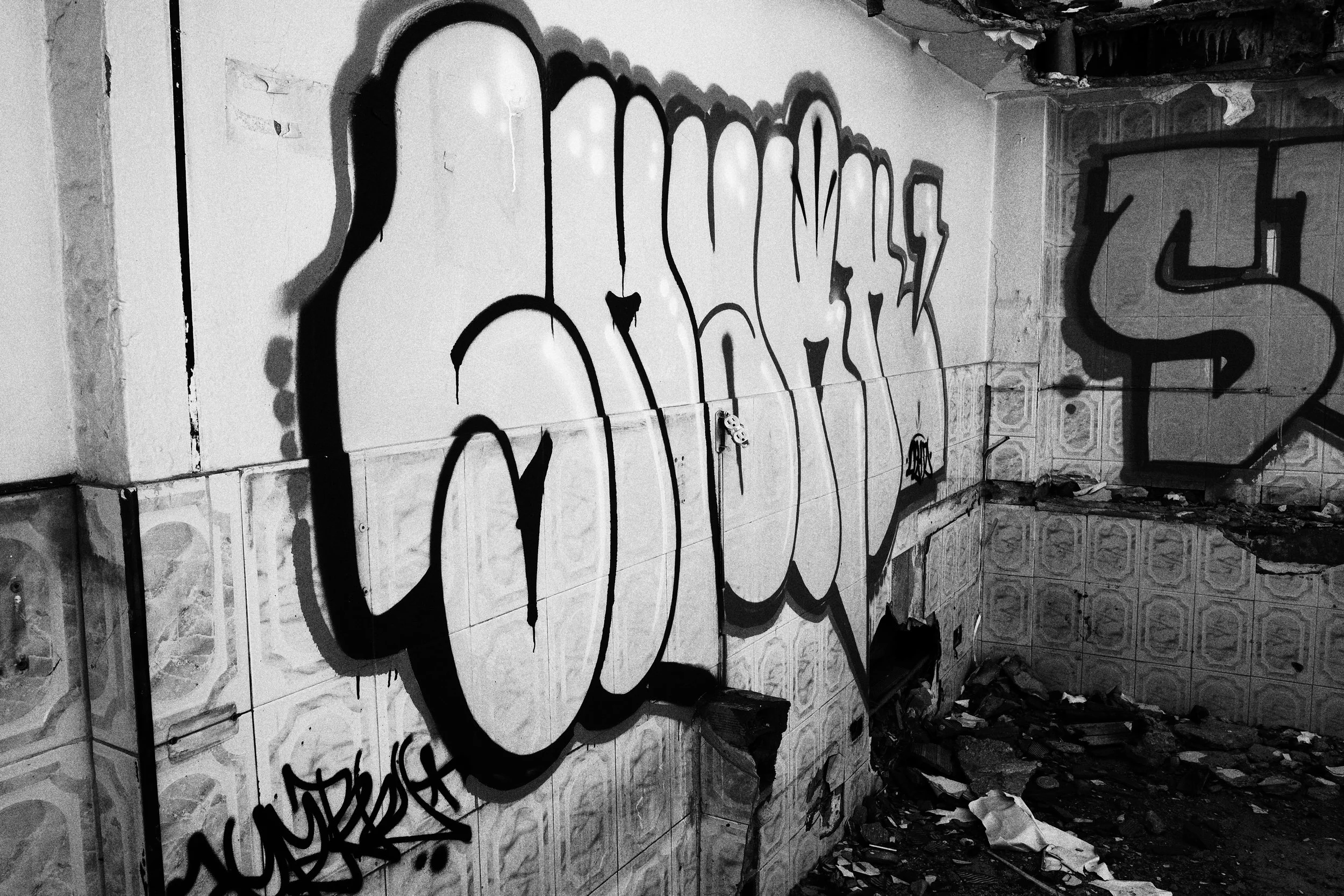 Black and white photo of large graffiti letters painted across tiled walls inside an abandoned room in Medellín, where debris and collapsed materials cover the dark floor.