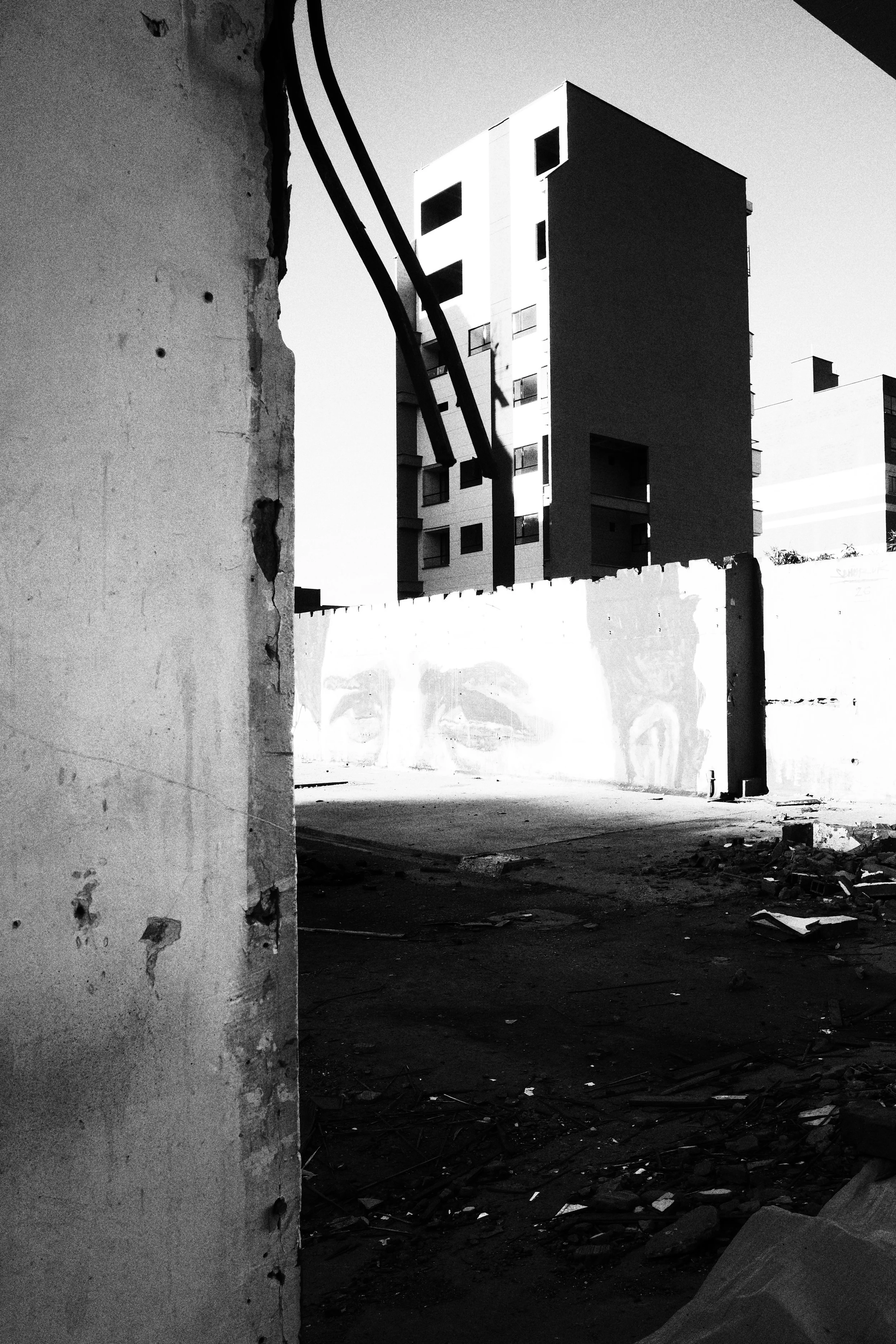 Black and white photo looking through the remains of a demolished structure in Medellín toward bright apartment towers, with exposed cables hanging over a nearly blank concrete wall.
