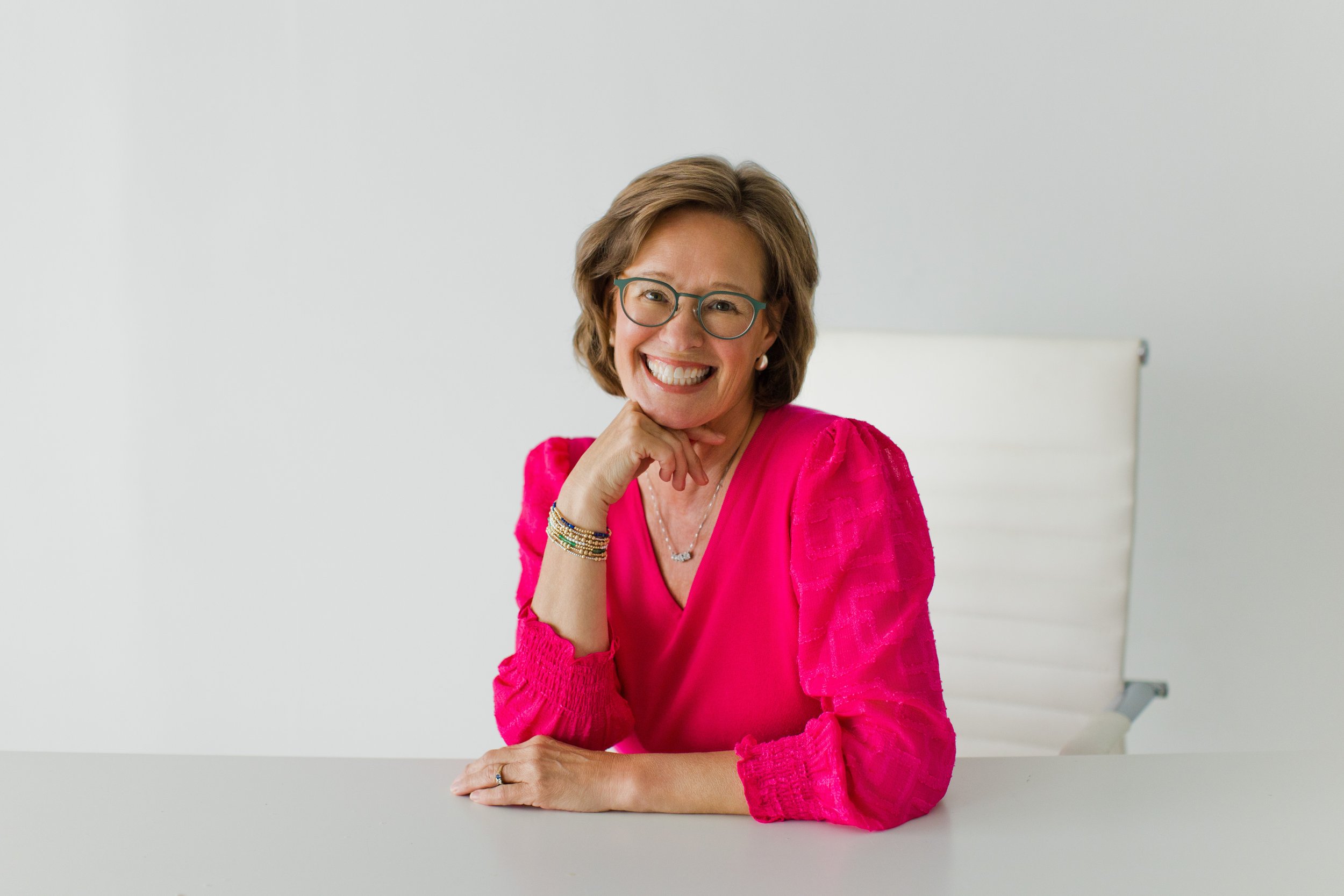smiling woman in a pink shirt with her hand under her chin sitting at a white desk with white walls behind her
