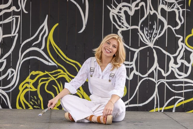 Artist lifestyle headshot of woman sitting in front of floral mural holding paintbrush by K Boyer Real Estate Media in Western North Carolina”