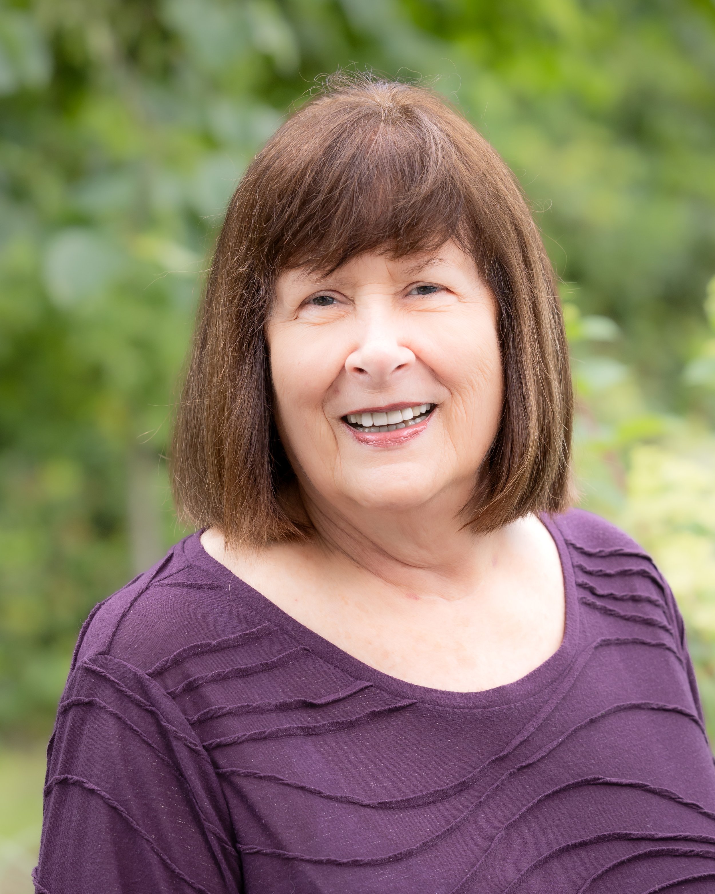 Headshot of a woman with short brown hair wearing a purple shirt smiling at the camera iwth a soft green backdrop of leaves and trees