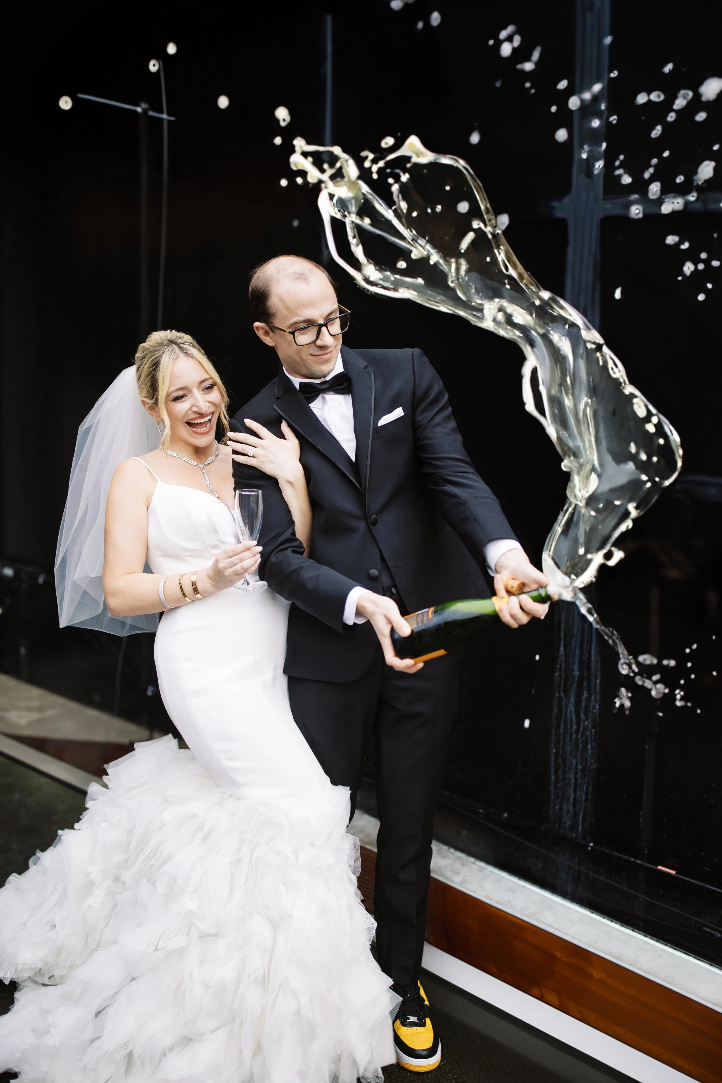 Bride and groom popping champagne, bride in white dress and groom in tuxedo with black and yellow shoes, champagne spraying.