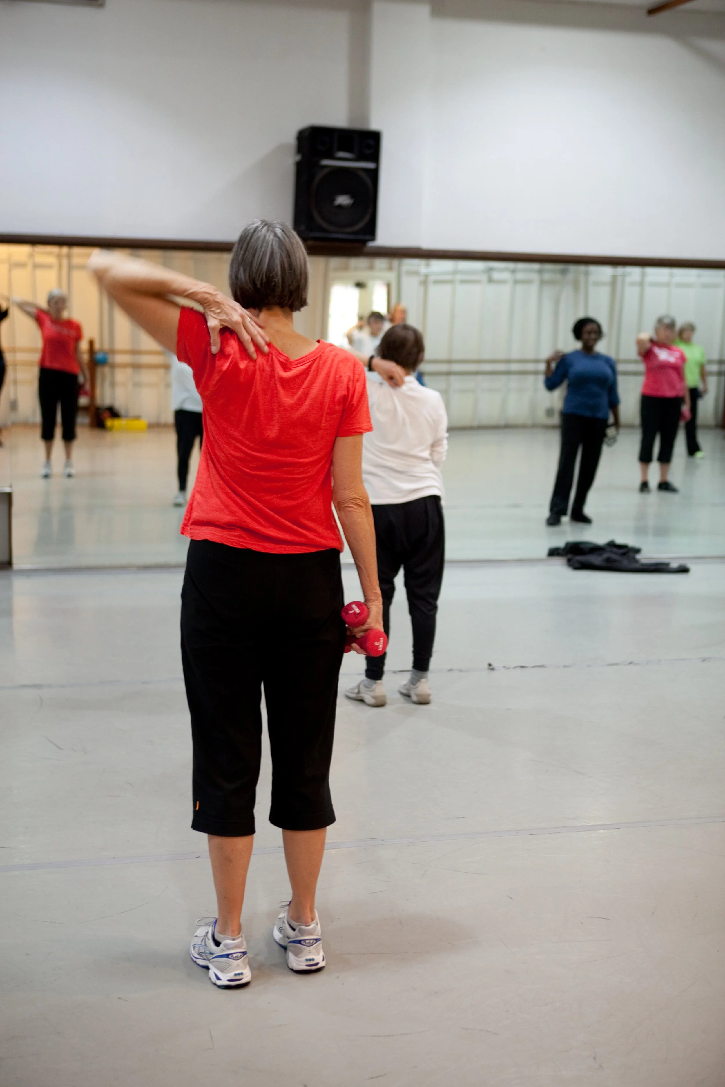 A group of older women participating in a fitness class in front of a mirror, with one woman in the foreground holding small red dumbbells and stretching her arm.
