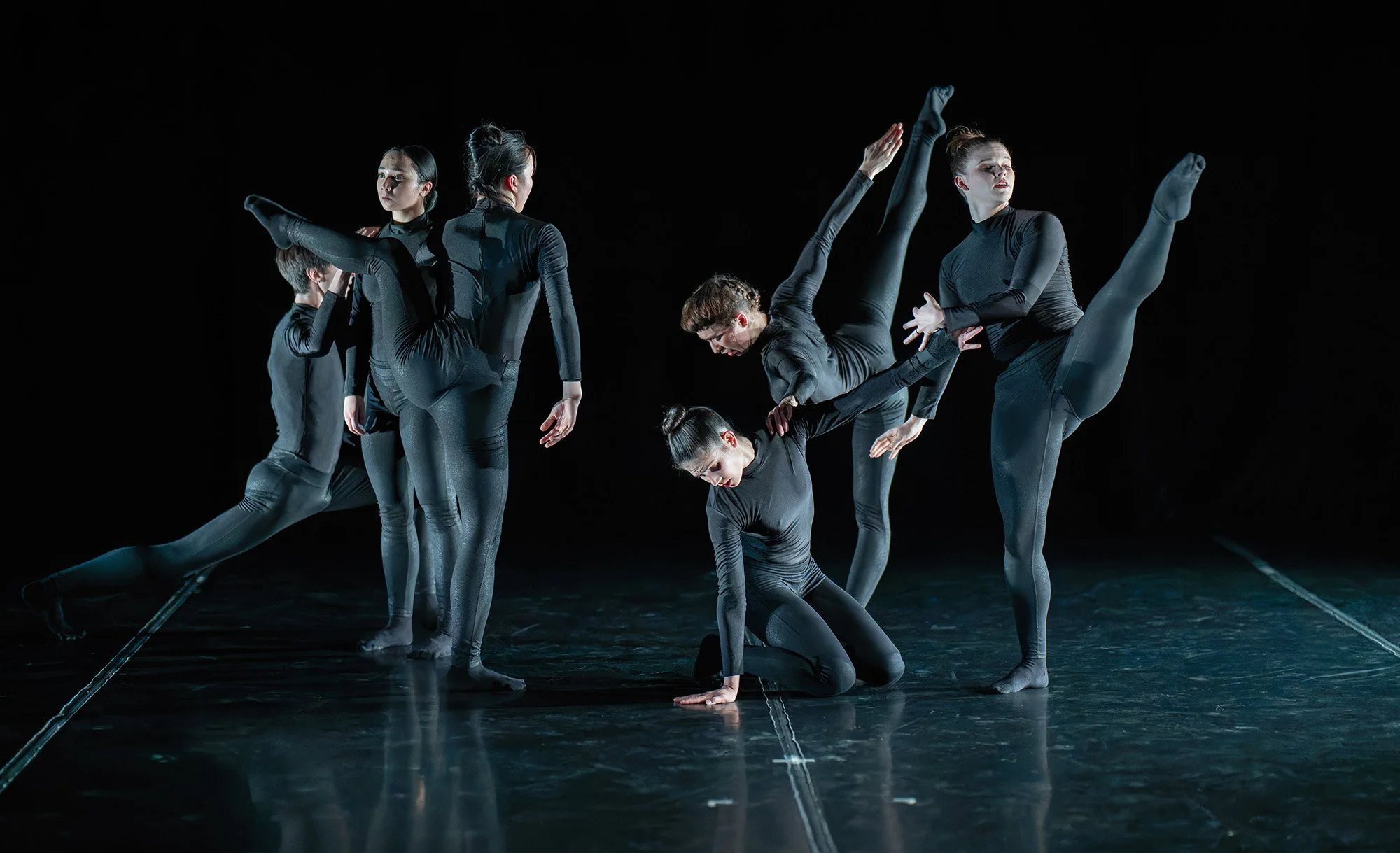 Group of six dancers in black costumes performing on dark stage, engaging in a contemporary dance routine with various expressive poses and movements.