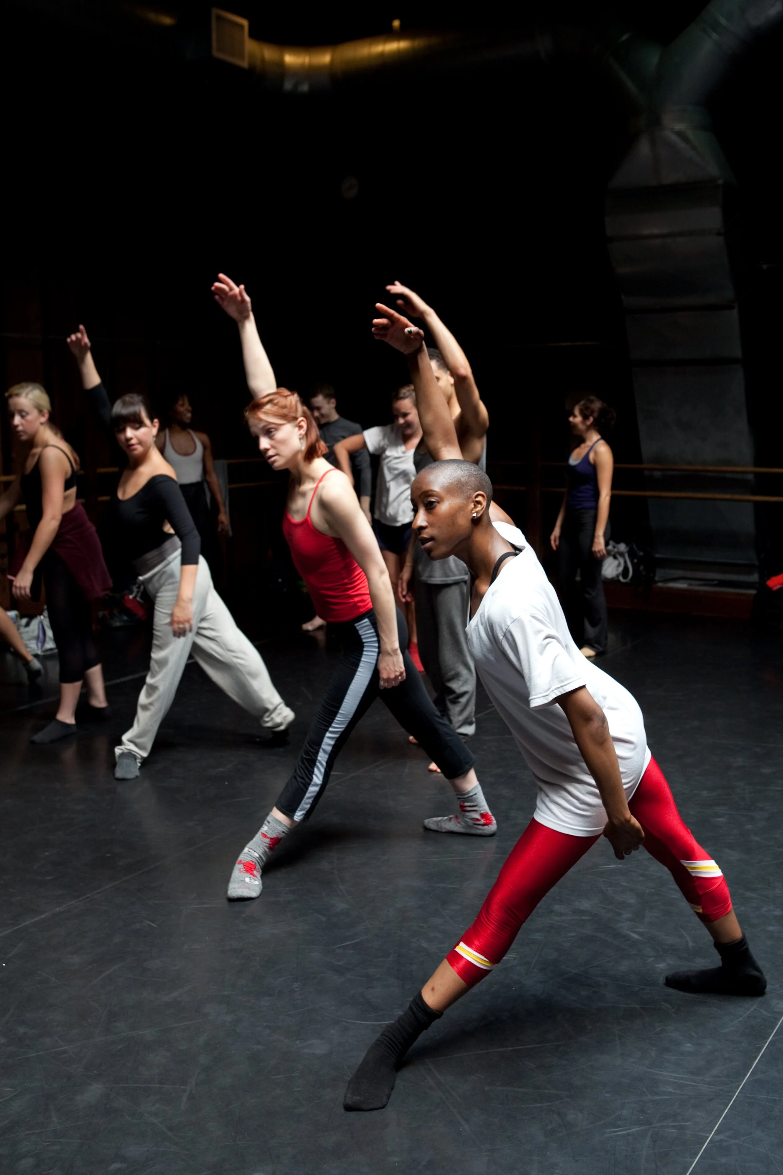 Group of diverse dancers practicing choreography in a dance studio with black floors and dark walls.