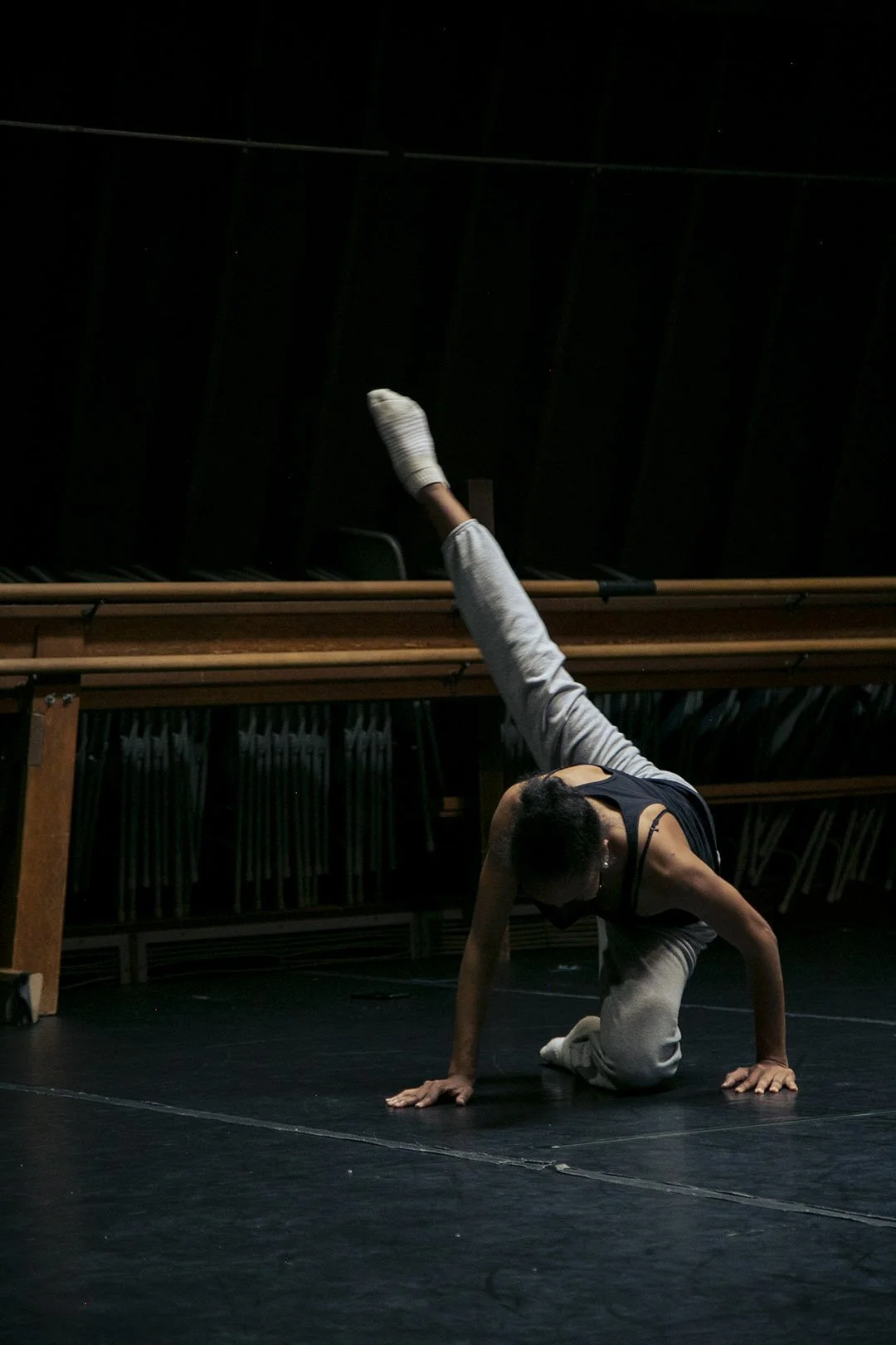 A dancer performs a move on stage, with one hand and knee on the ground, one leg extended upward, dressed in grey sweatpants and a black tank top, in a dark room with stage lighting.