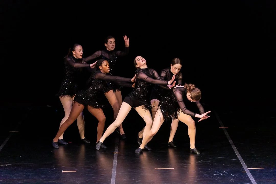 Six young female dancers in black costumes perform on stage with a dark background.