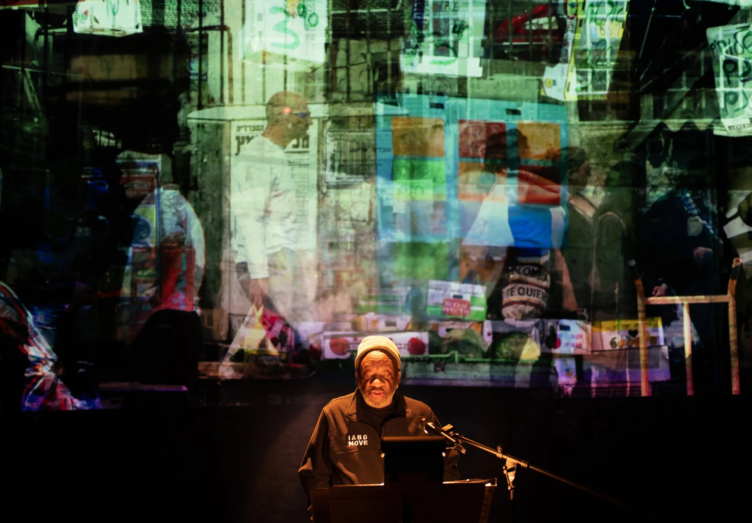 An elderly man with a beard and yellow knit cap standing at a keyboard with a microphone, performing on stage. Behind him, there is a large, colorful, layered projection of a busy marketplace scene with people shopping and signs, creating an abstract, multi-dimensional background.