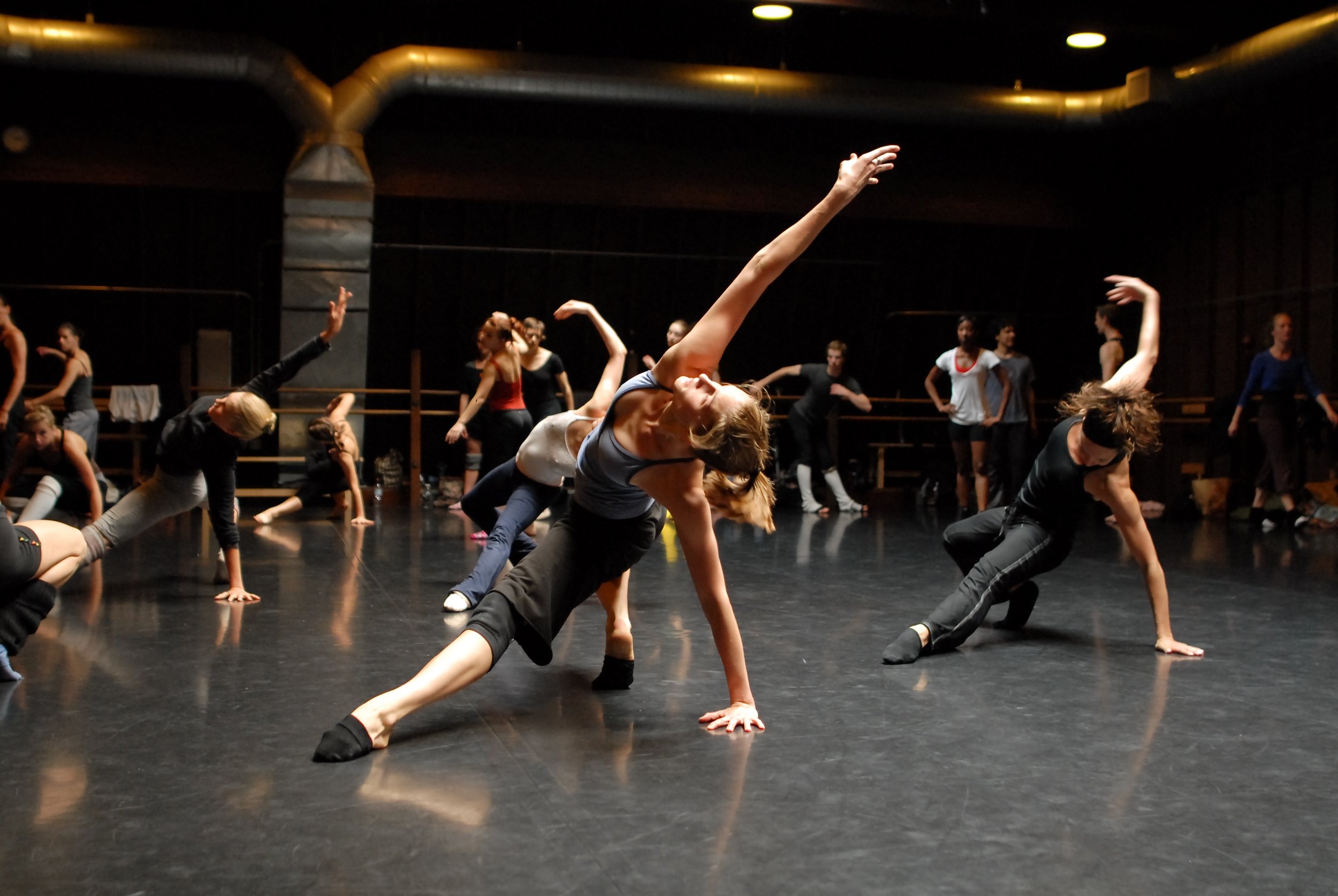 A group of dancers practicing contemporary dance movements on a dark studio floor, with some dancers in the background stretching or warming up.