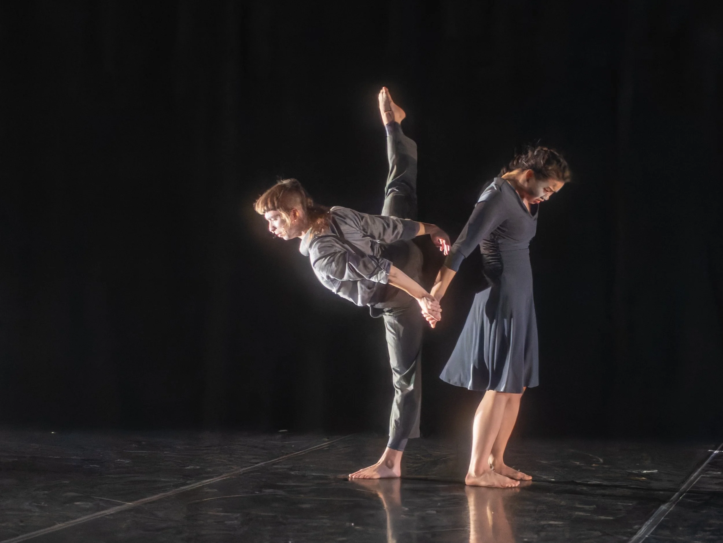 Two female contemporary dancers perform on stage, one supporting the other, who is balanced on one leg with the other extended upward. They hold hands, dressed in dark, flowy costumes, with a black background and spotlight highlighting them.