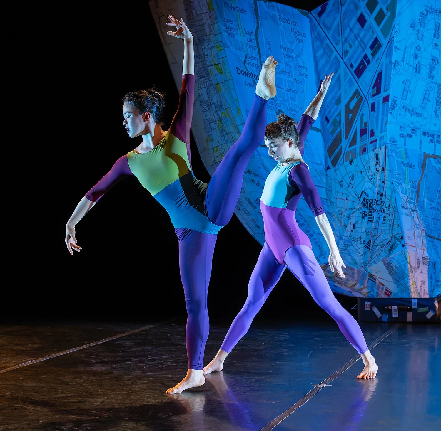 Two dancers performing on stage in colorful bodysuits, with a map of a city in the background.