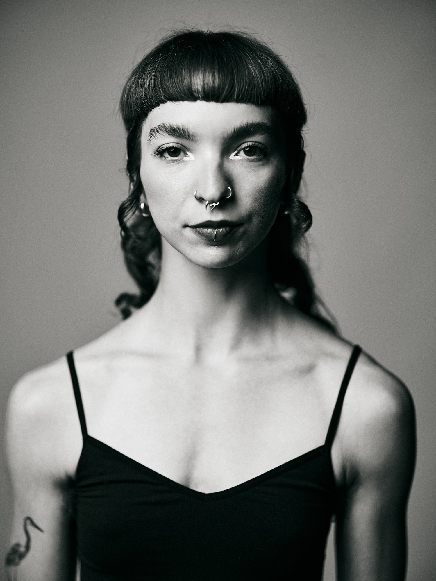 Black and white portrait of a woman with short dark hair, bangs, multiple facial piercings, wearing a spaghetti strap top, with a tattoo of a bird on her right arm.