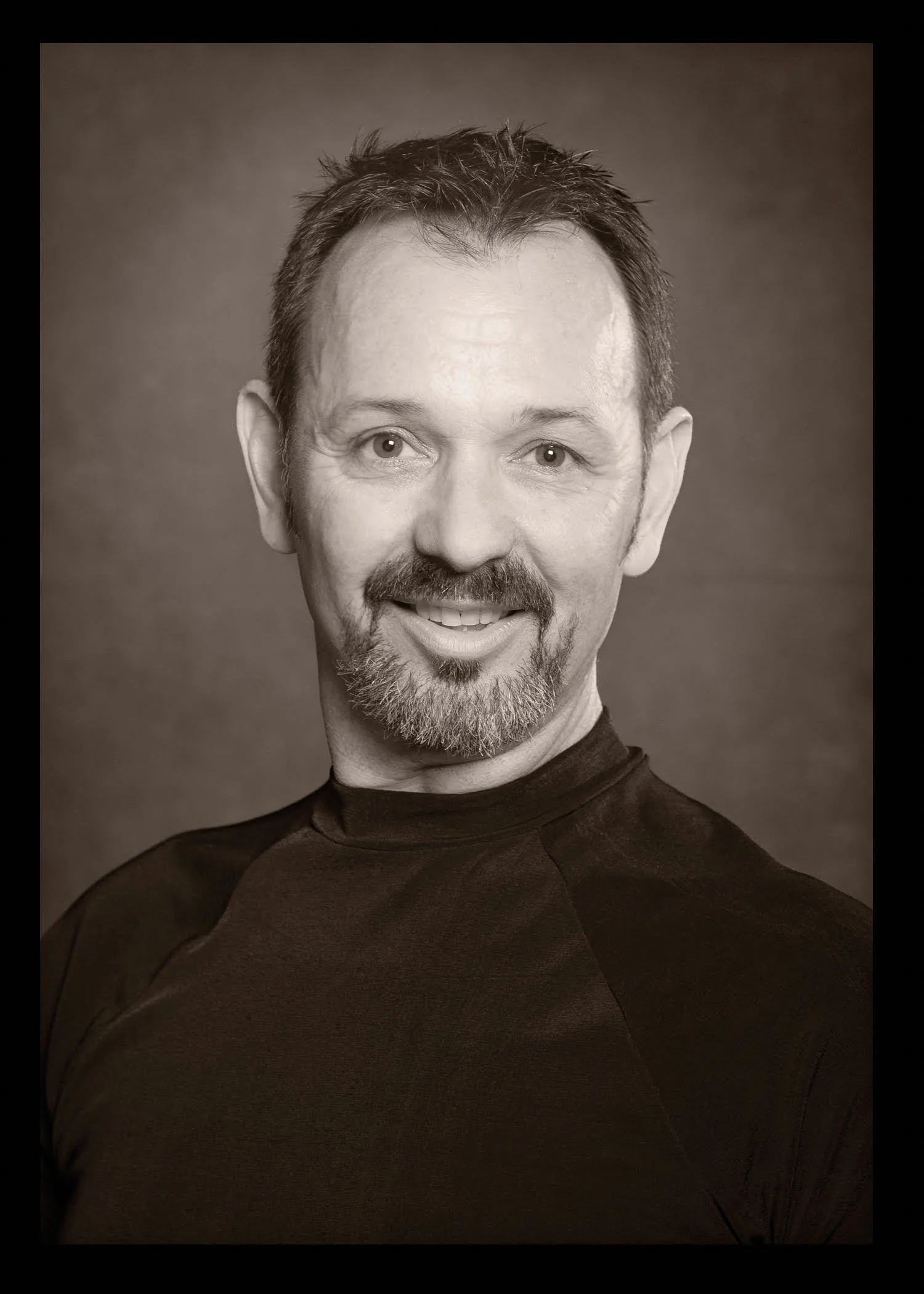 A black and white portrait of a smiling man with short hair, a beard, and mustache, wearing a dark athletic shirt, against a plain background.