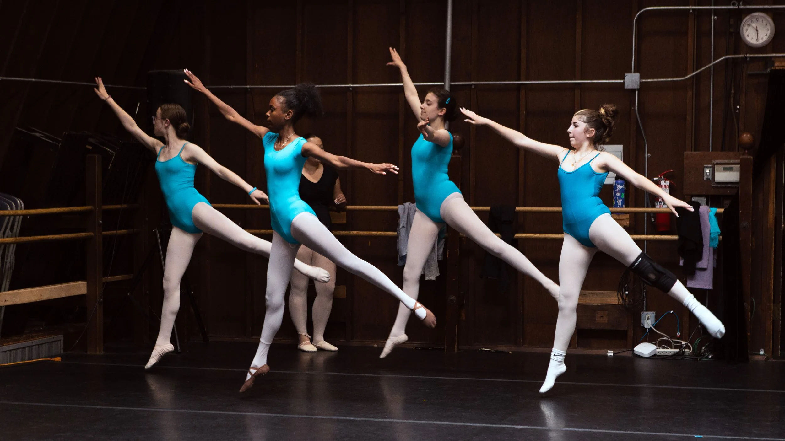 Group of young ballet dancers practicing in a dance studio, wearing blue leotards and white tights, performing jumps near a wooden ballet barre.