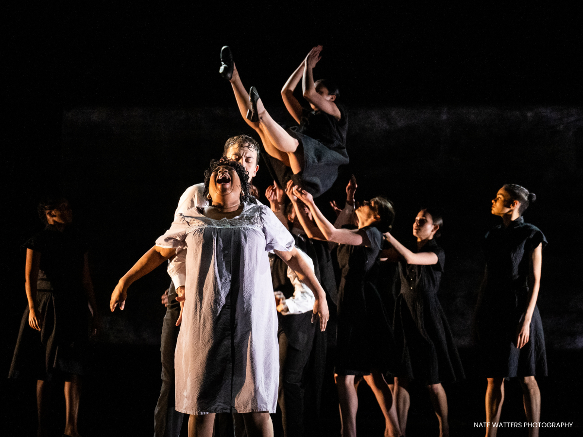 A theatrical dance performance featuring multiple performers dressed in black, with one woman in a white dress, lifting another performer. The performers are on a dark stage with dramatic lighting.