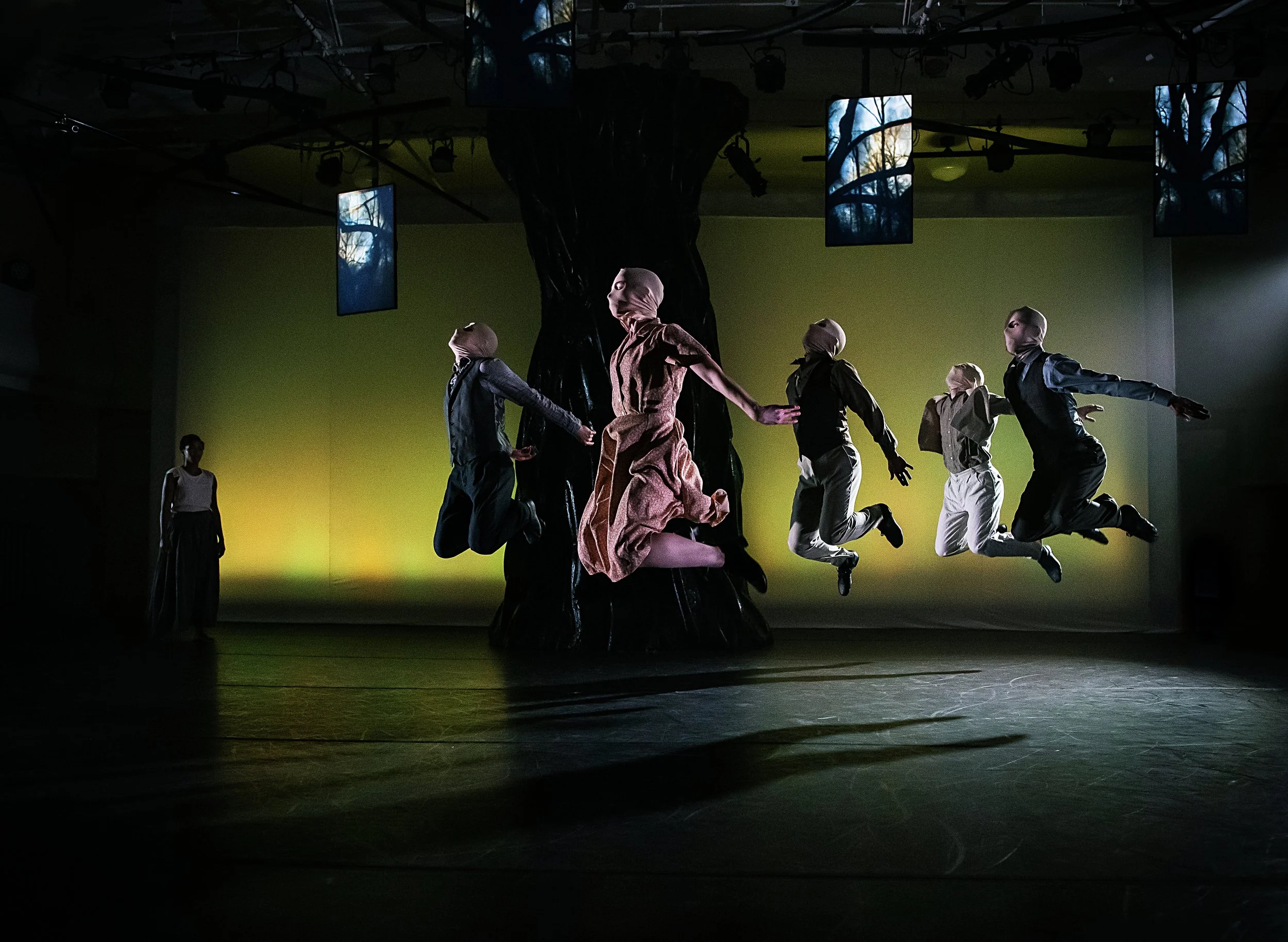 A theatrical dance performance with six dancers leaping in front of a large tree prop on stage with a yellow backdrop and digital screens showing images of trees above.
