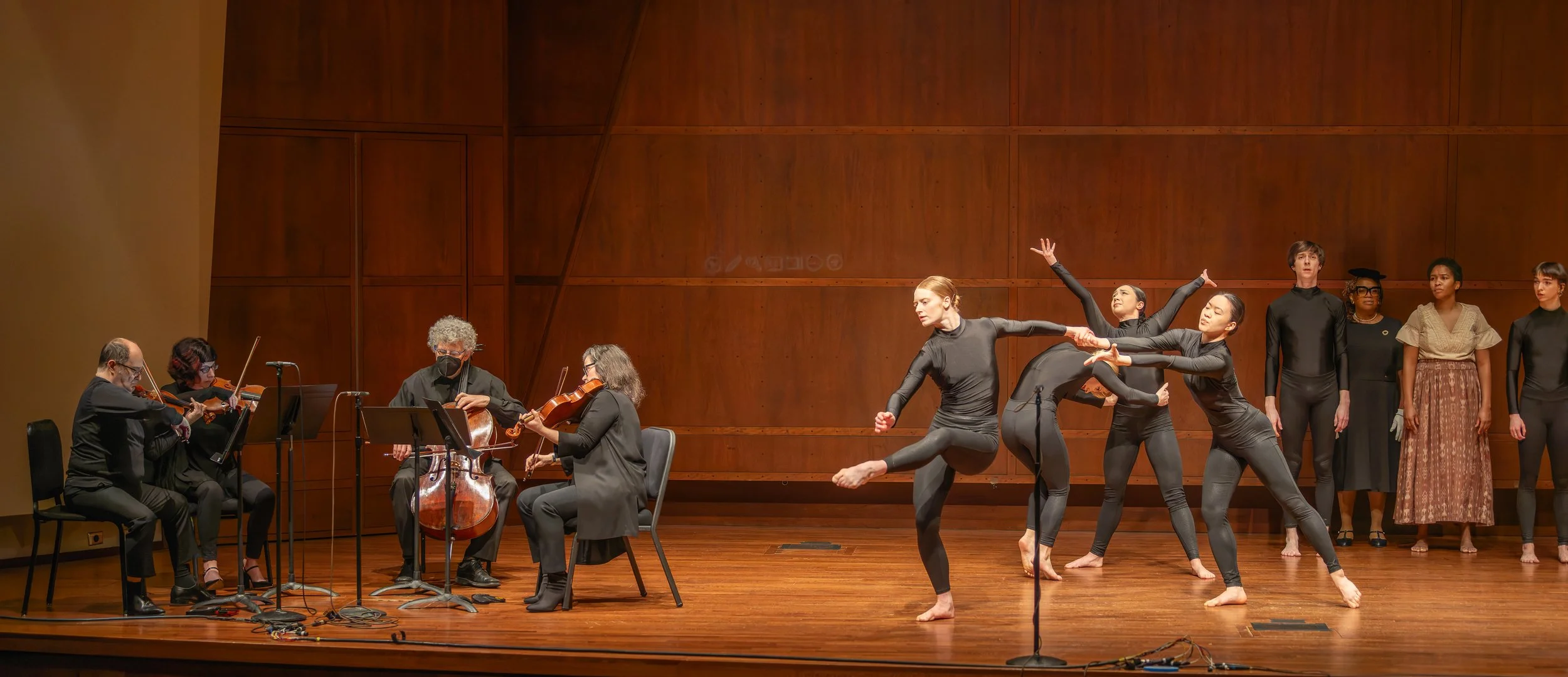 A group of ballet dancers in black leotards performing on stage, with an orchestra of string musicians playing nearby. The stage has a wooden floor and a wooden back wall.