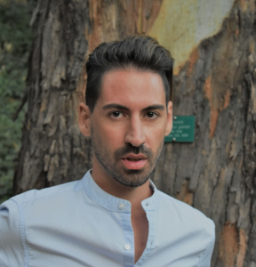 A man with dark hair and facial hair, wearing a light blue shirt, standing outdoors in front of a large tree.