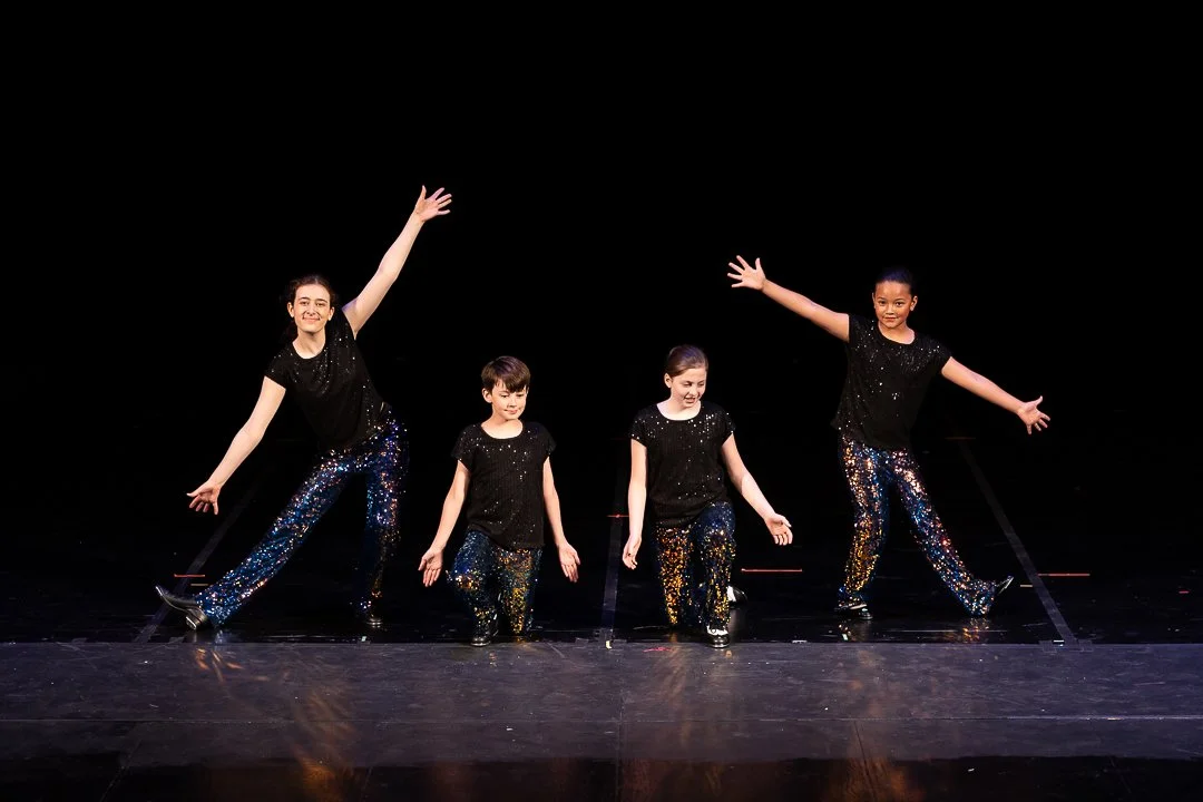Four young girls in black sequined shirts and matching pants performing a dance on stage with a black background, with the two on the ends raising one arm and the two in the middle kneeling.