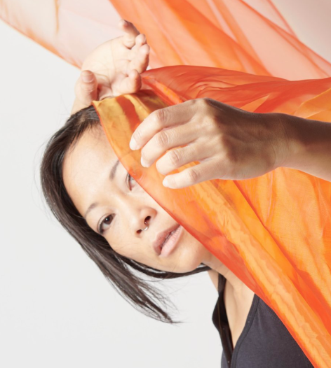 A woman adjusting orange and red translucent fabric, with a neutral background.