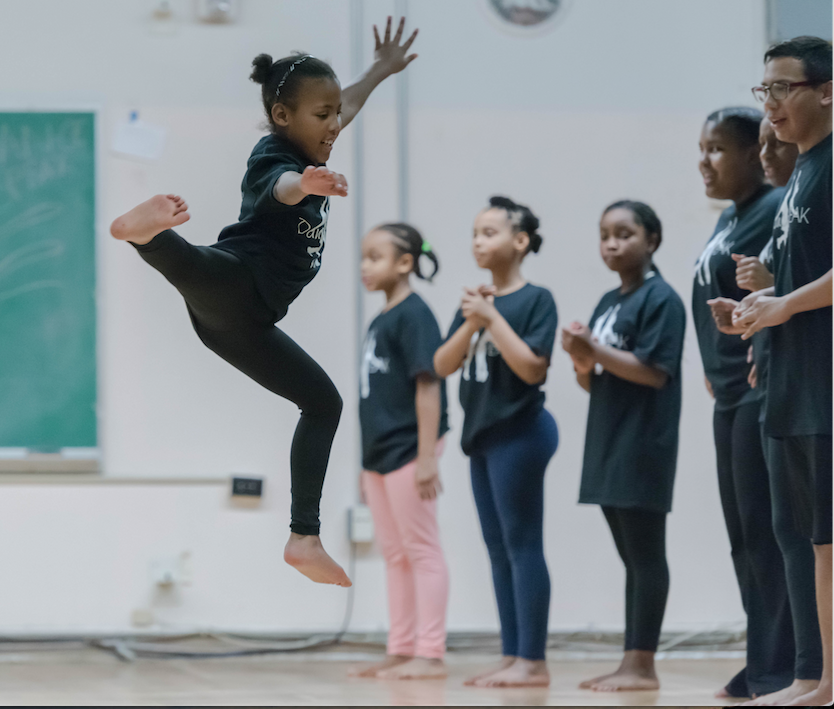 Young girl jumping in front of a group of children and a teacher in a classroom.