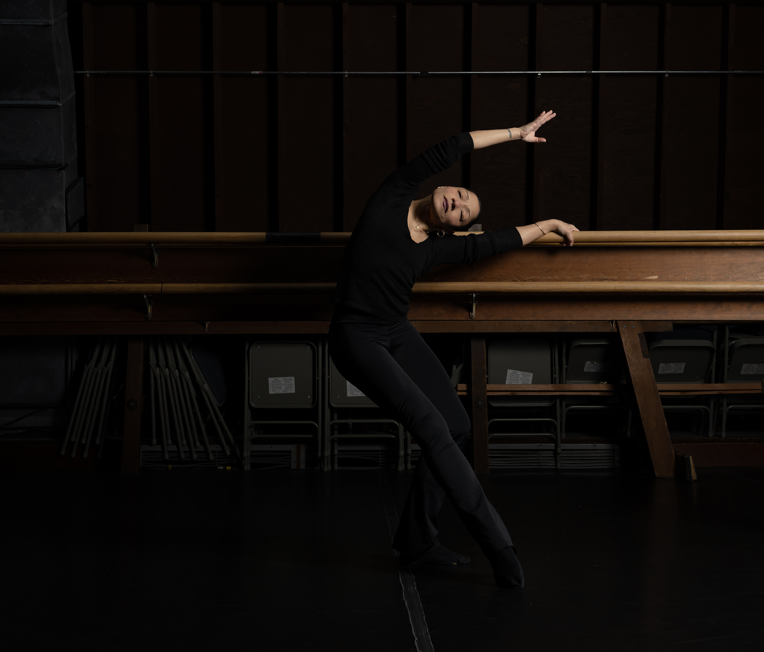 A woman in black dance attire poses in front of a ballet barre in a dimly lit dance studio.