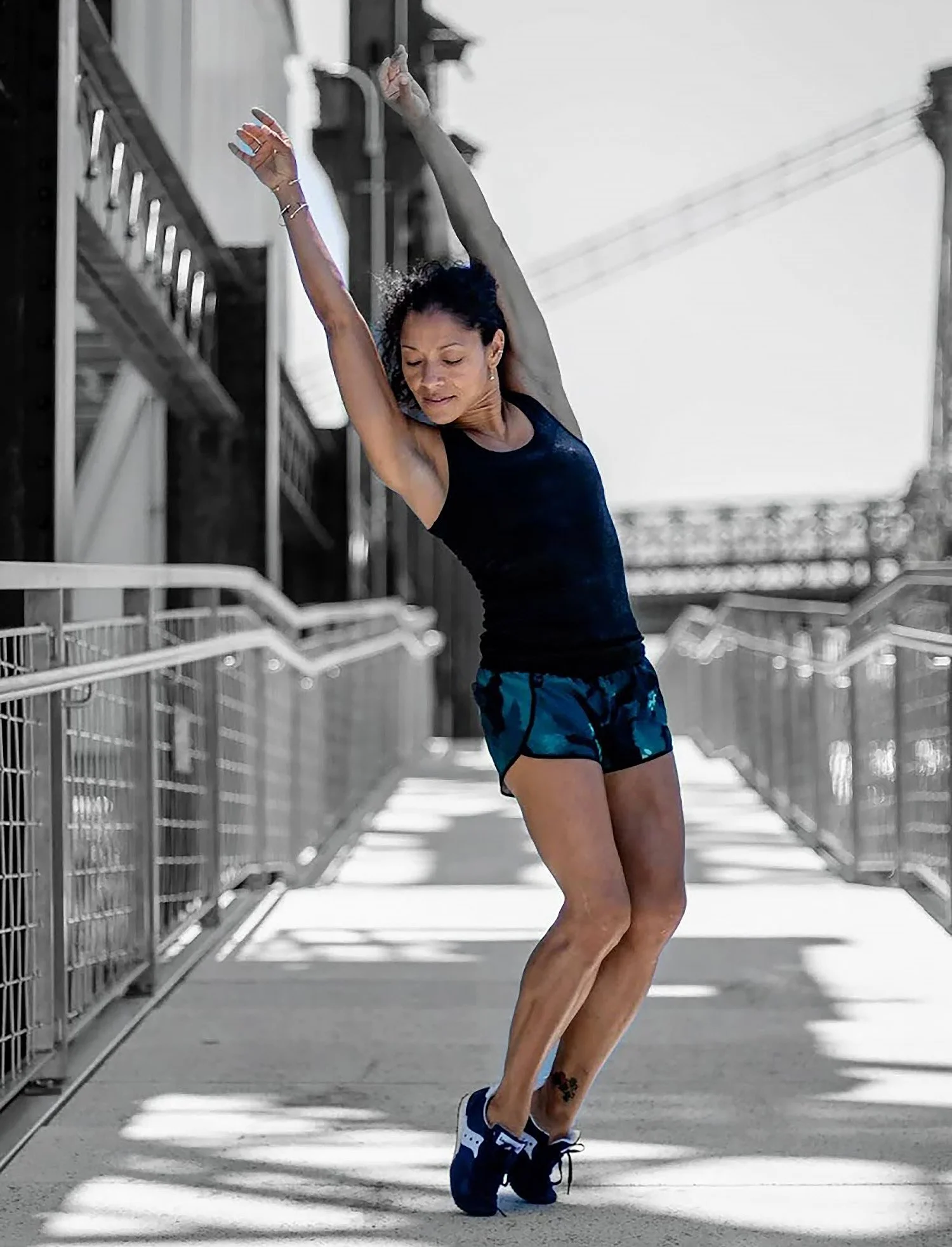 A woman with dark hair in a ponytail, wearing a black sleeveless top and blue athletic shorts, is stretching outdoors on a bridge, raising her arms above her head and bending to one side."